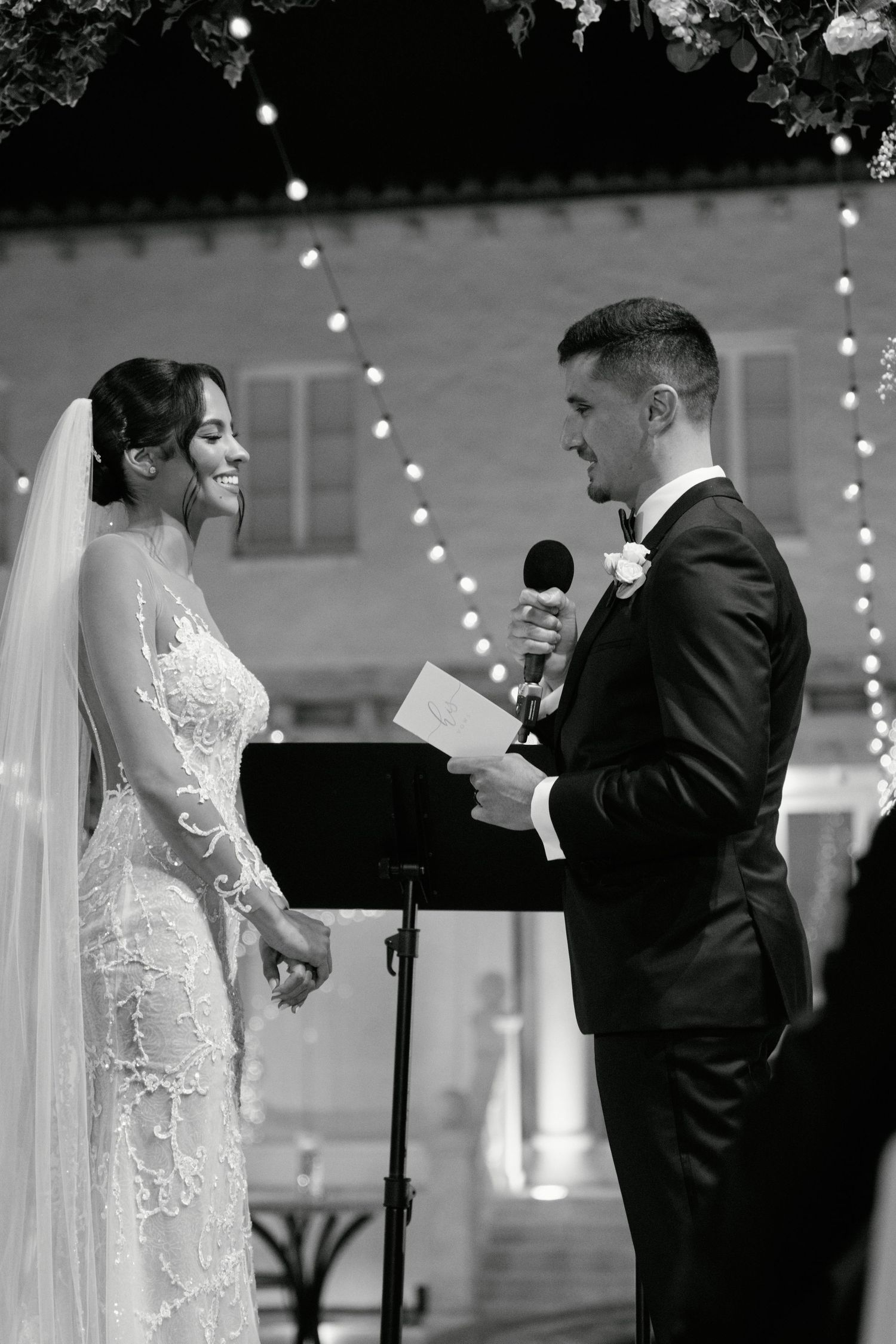 Black and white photo of wedding ceremony with couple exchanging vows under string lights.