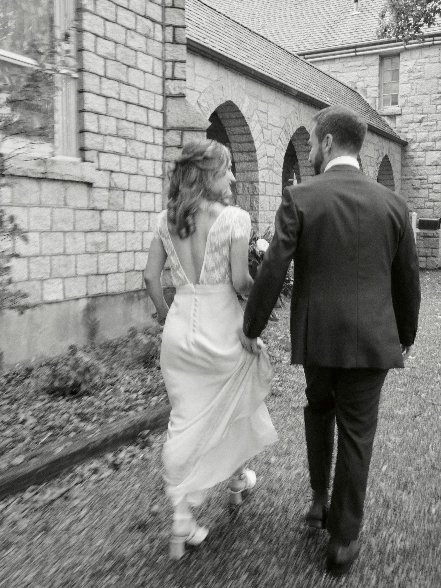 A couple in wedding attire walks along a stone church pathway in black and white.