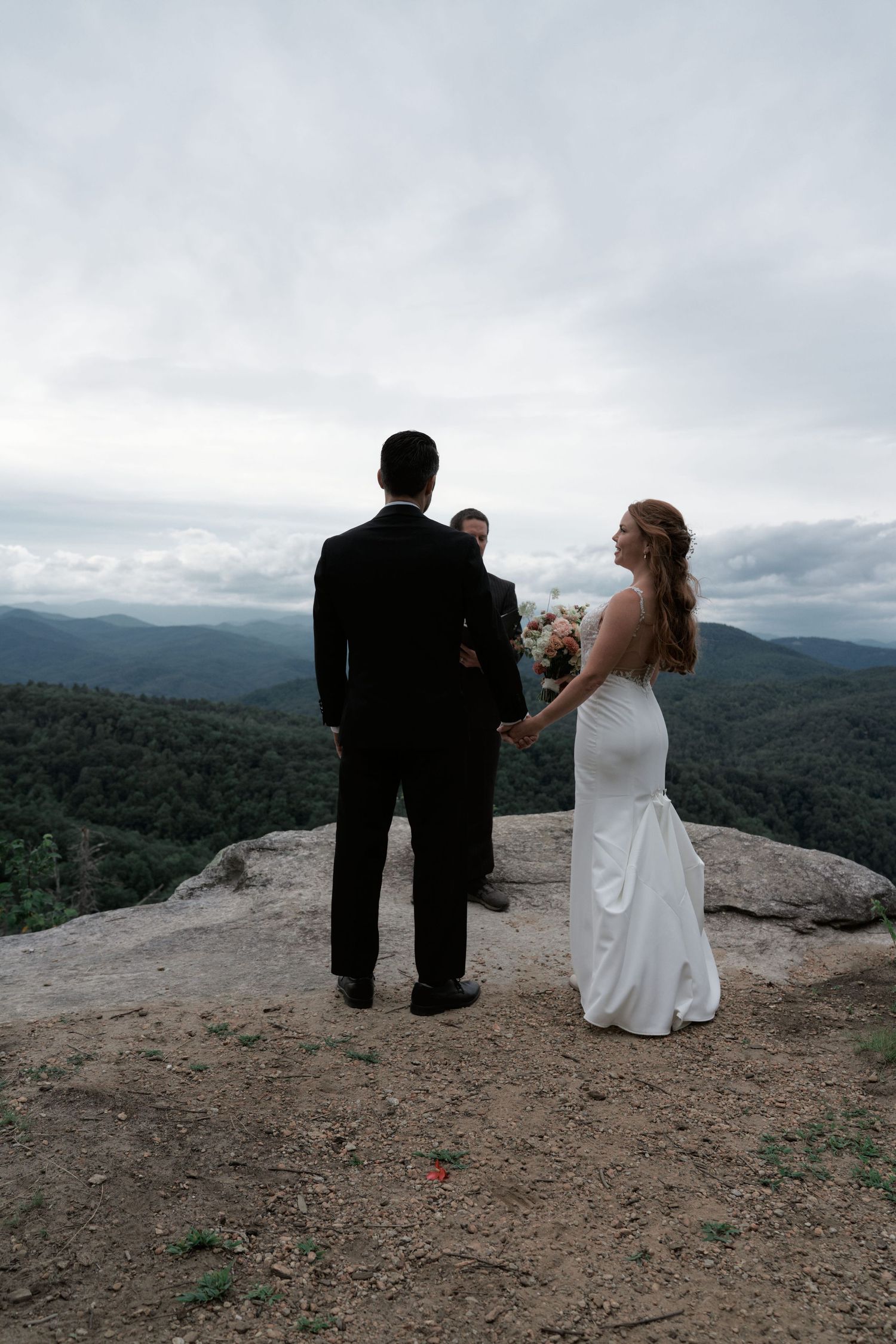 A couple in wedding attire stands at the edge of a mountain overlook with sweeping views of forested peaks below.