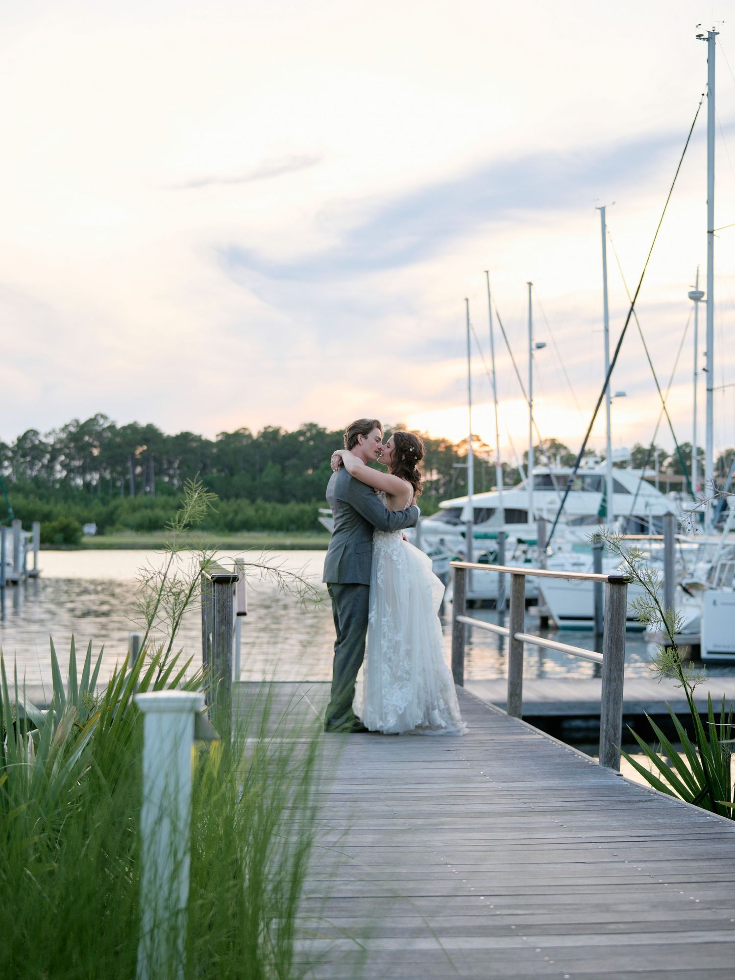 A romantic couple embraces on a wooden dock at sunset with boats and a marina in the background.
