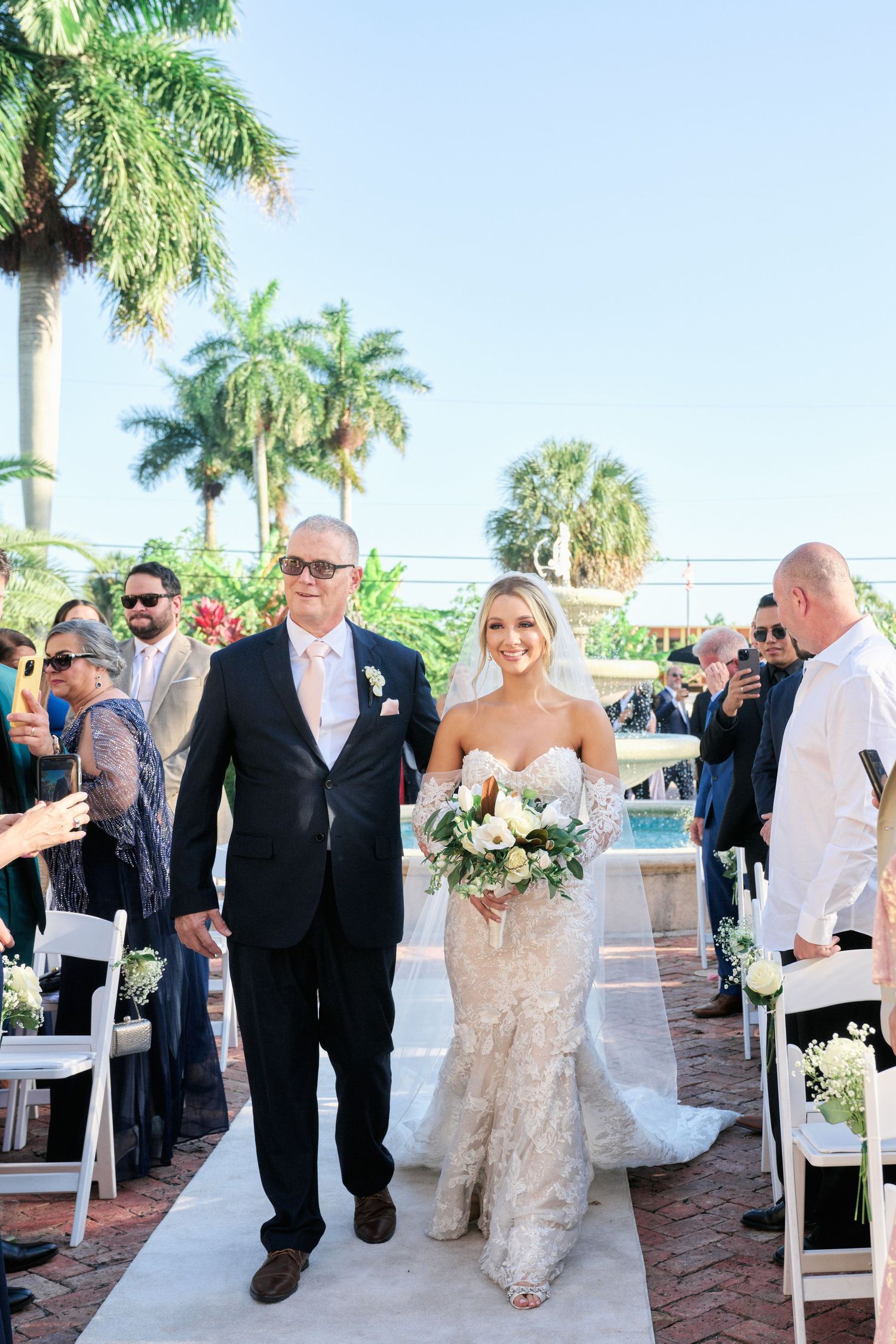 A wedding ceremony processional taking place on a palm-lined pathway with guests seated on white chairs.