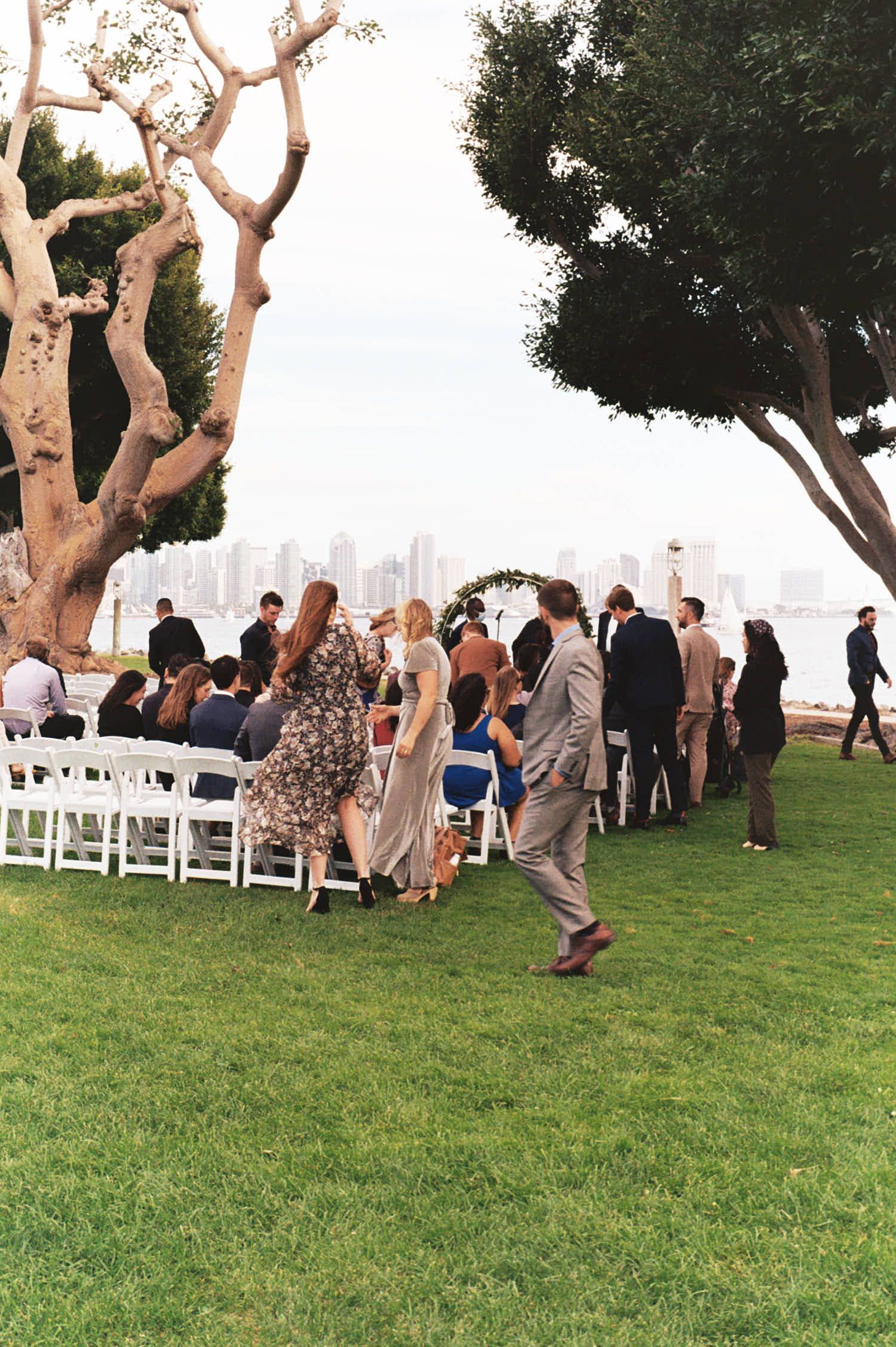 Outdoor wedding ceremony setup with guests gathering under large tree with city skyline in background.