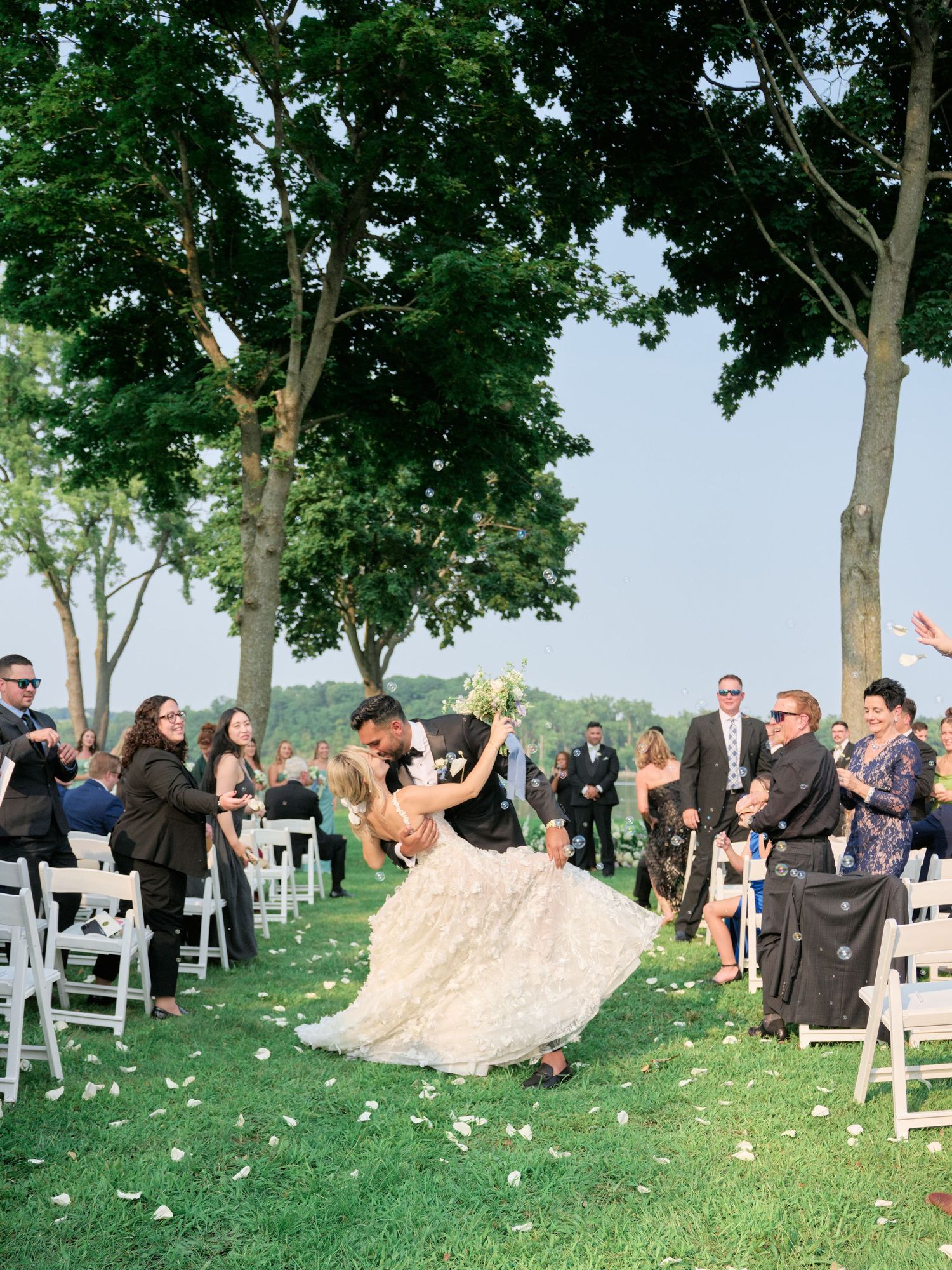 Outdoor wedding celebration under tall trees with guests seated in white chairs on a grassy lawn.