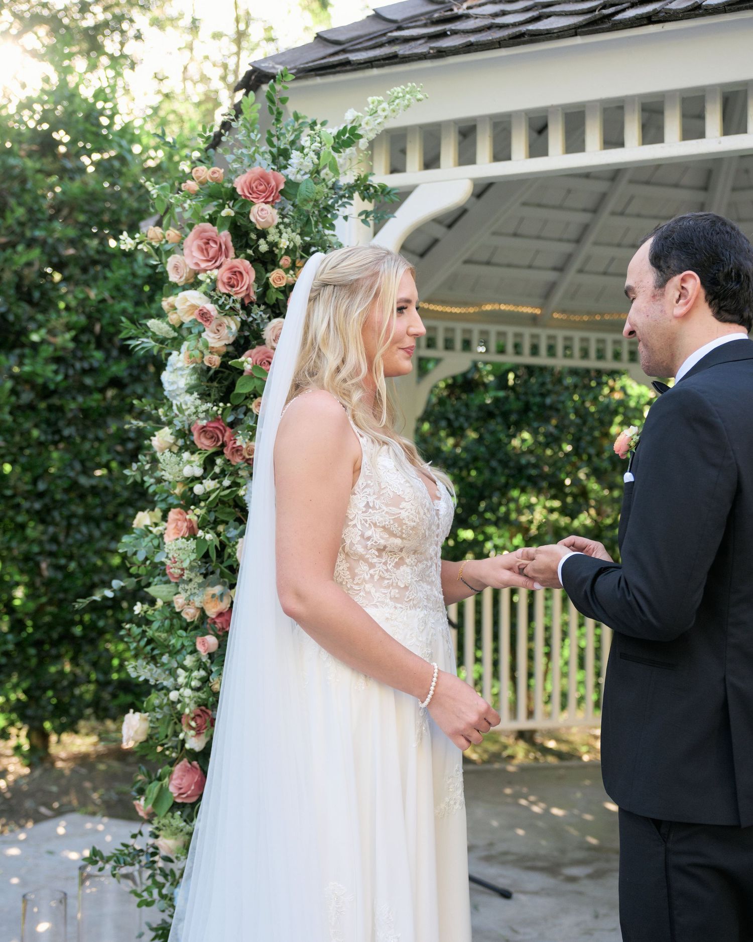 Wedding couple exchanges vows under a floral arch with pink roses in a garden setting.