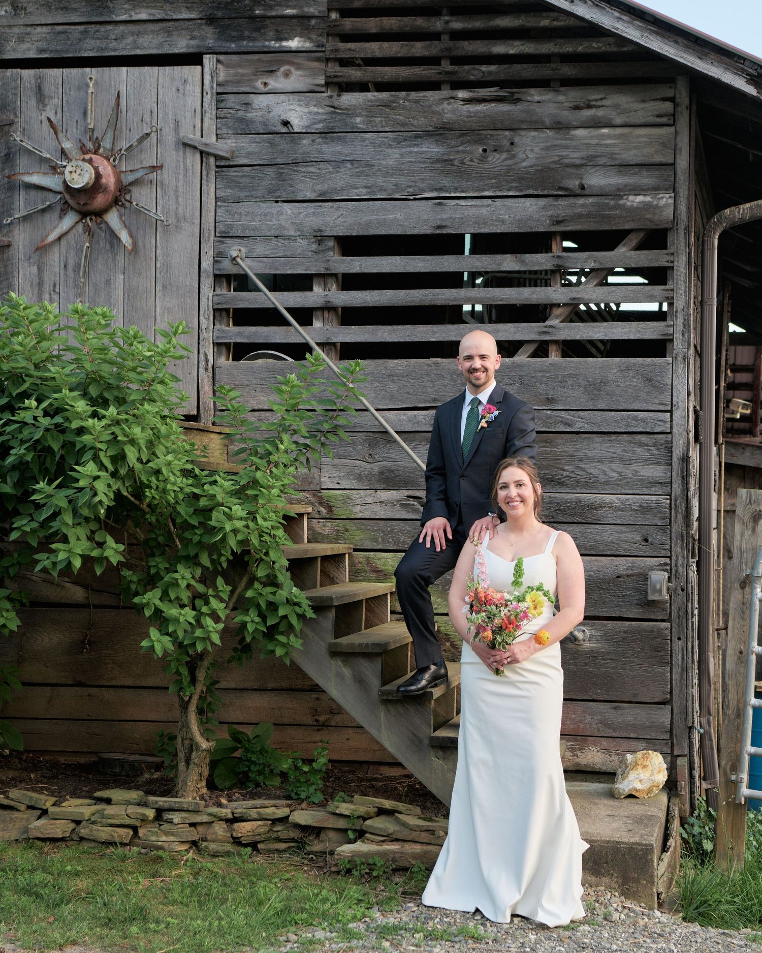 A couple poses on wooden stairs against a rustic barn backdrop holding a vibrant bouquet.