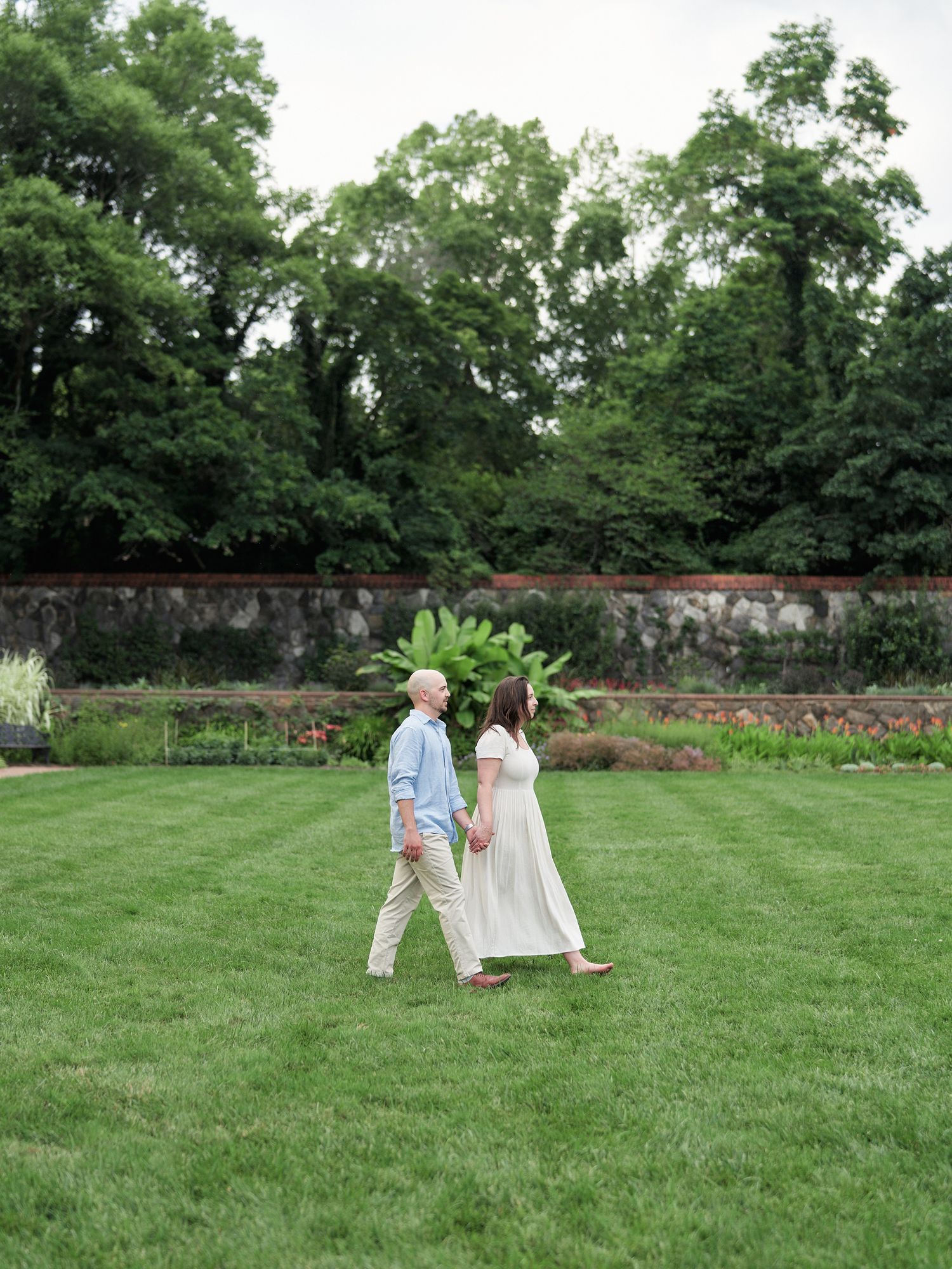 Two people walk together on a green lawn with stone walls and trees creating a romantic garden setting.