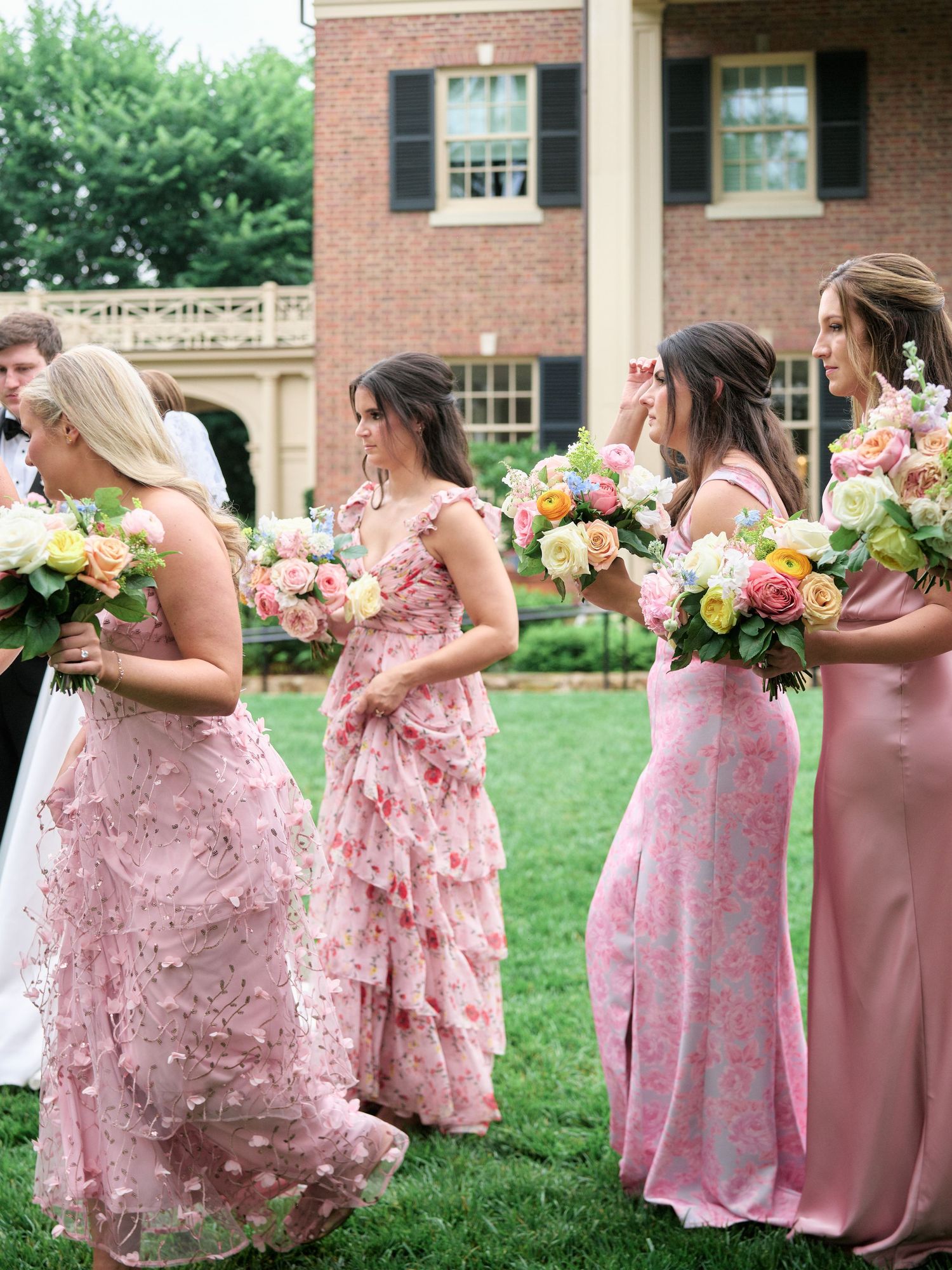 Bridesmaids in varied pink dresses hold colorful bouquets on mansion lawn.