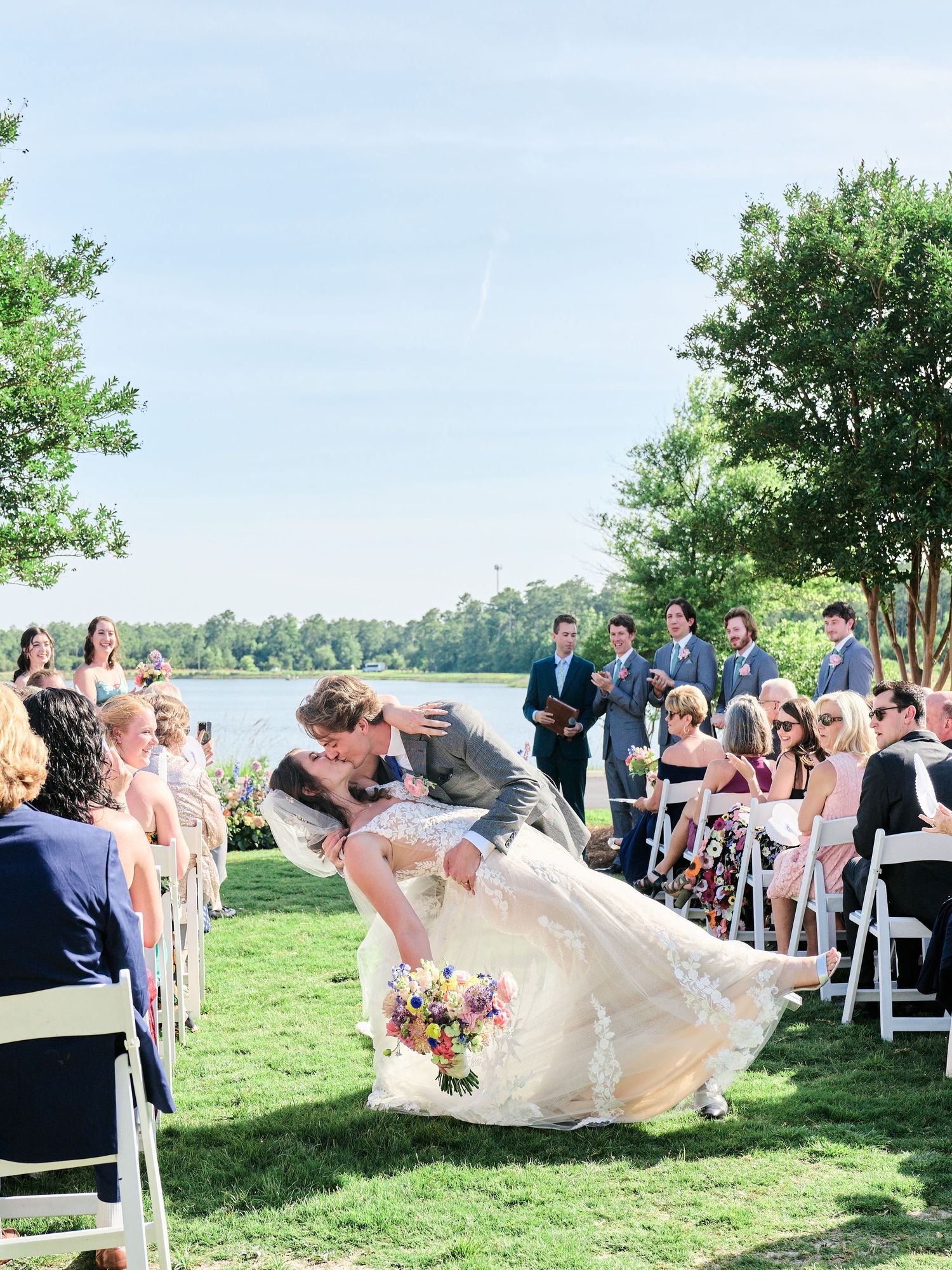 A romantic outdoor wedding ceremony by a lake where a couple kisses while the wind dramatically lifts the bride's dress.