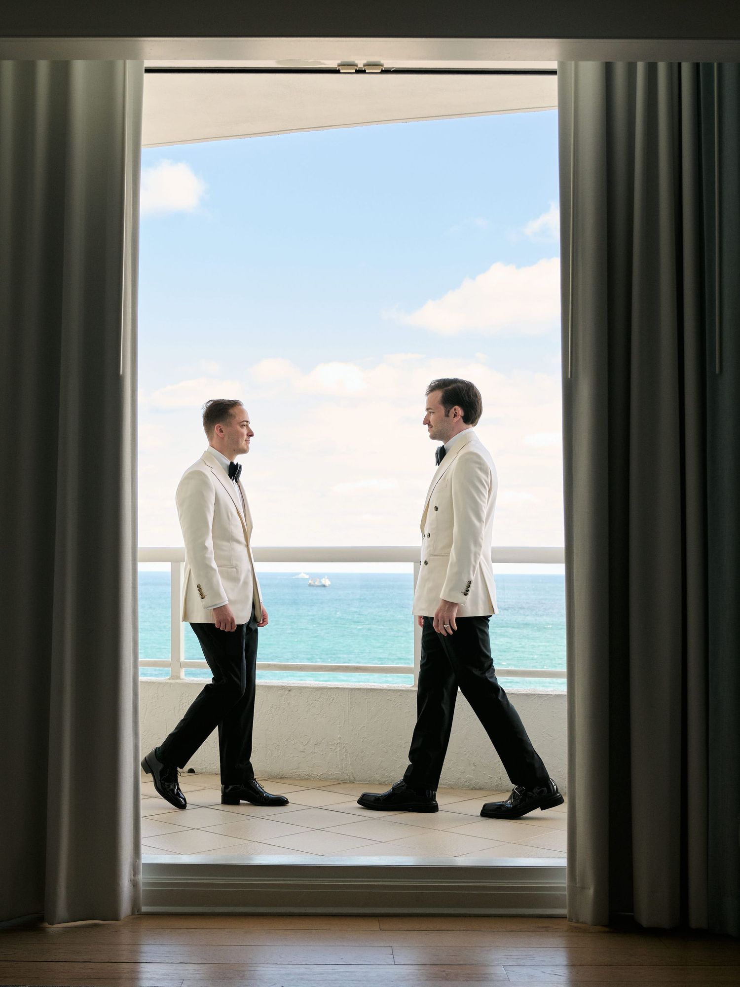 Two people in white jackets stand facing each other on an oceanfront balcony framed by curtains.