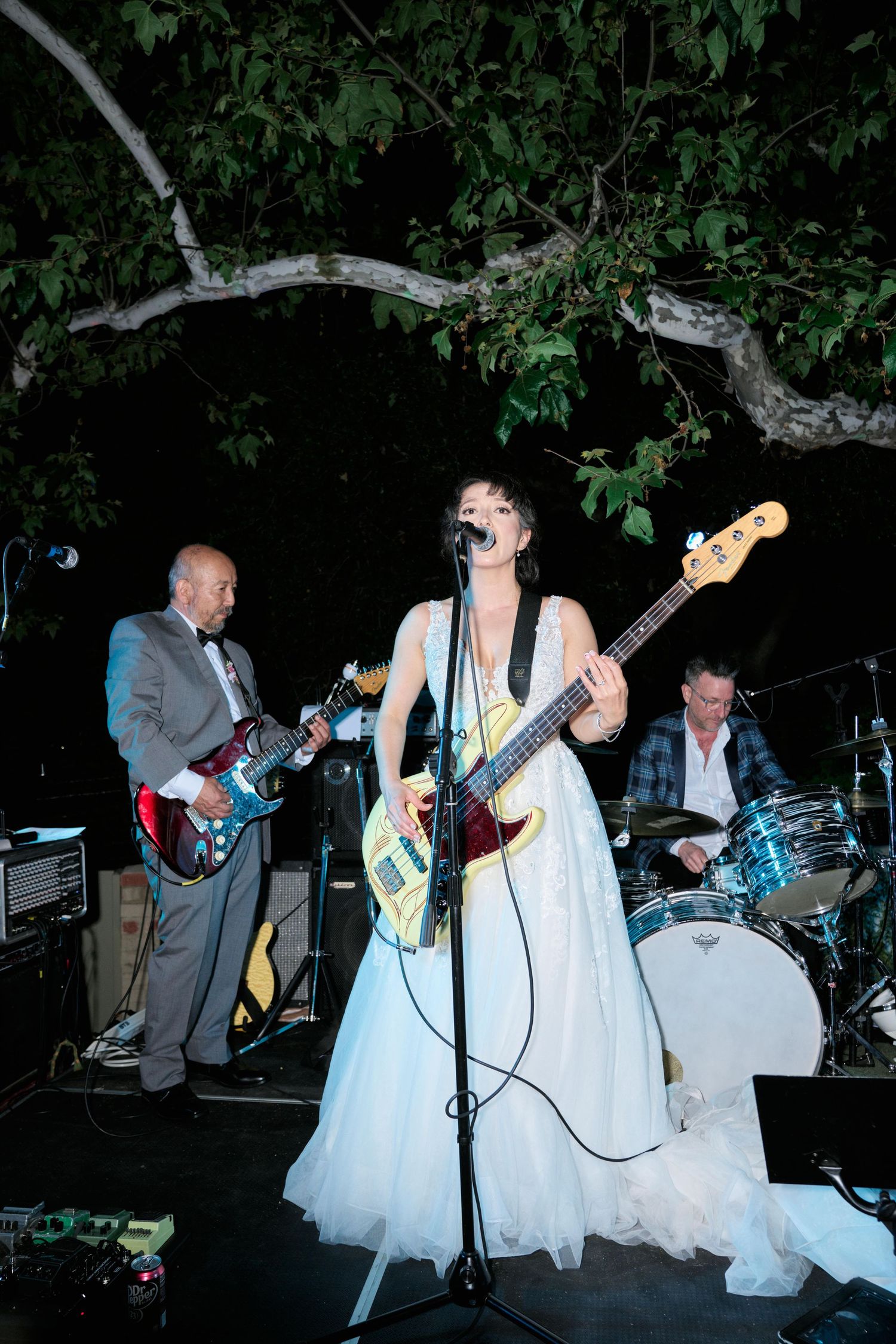 Musicians performing outdoors at night with white-dressed performer playing bass guitar.