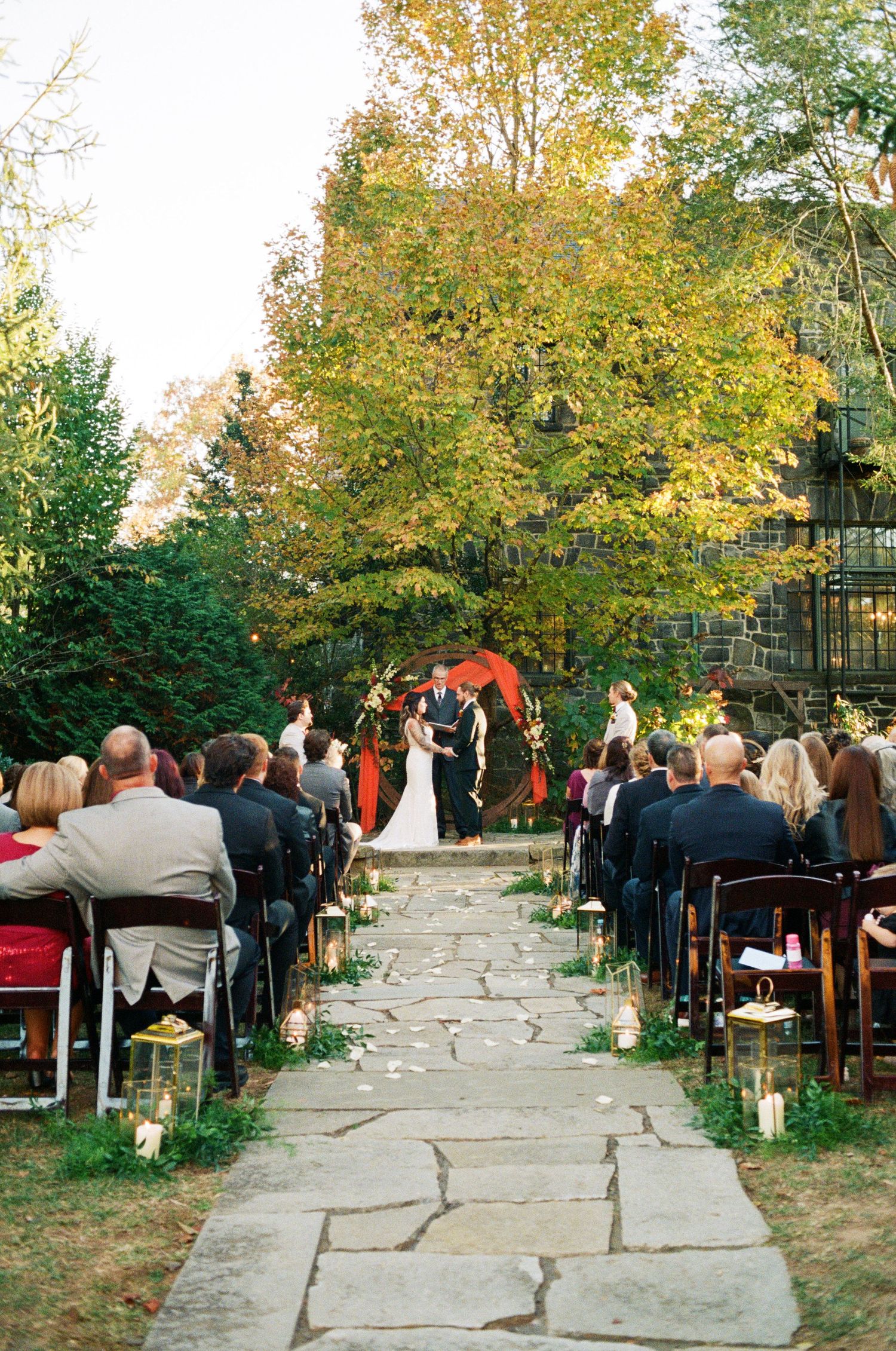 Guests seated outdoors at a fall wedding ceremony with autumn foliage and stone pathway leading to the altar area.