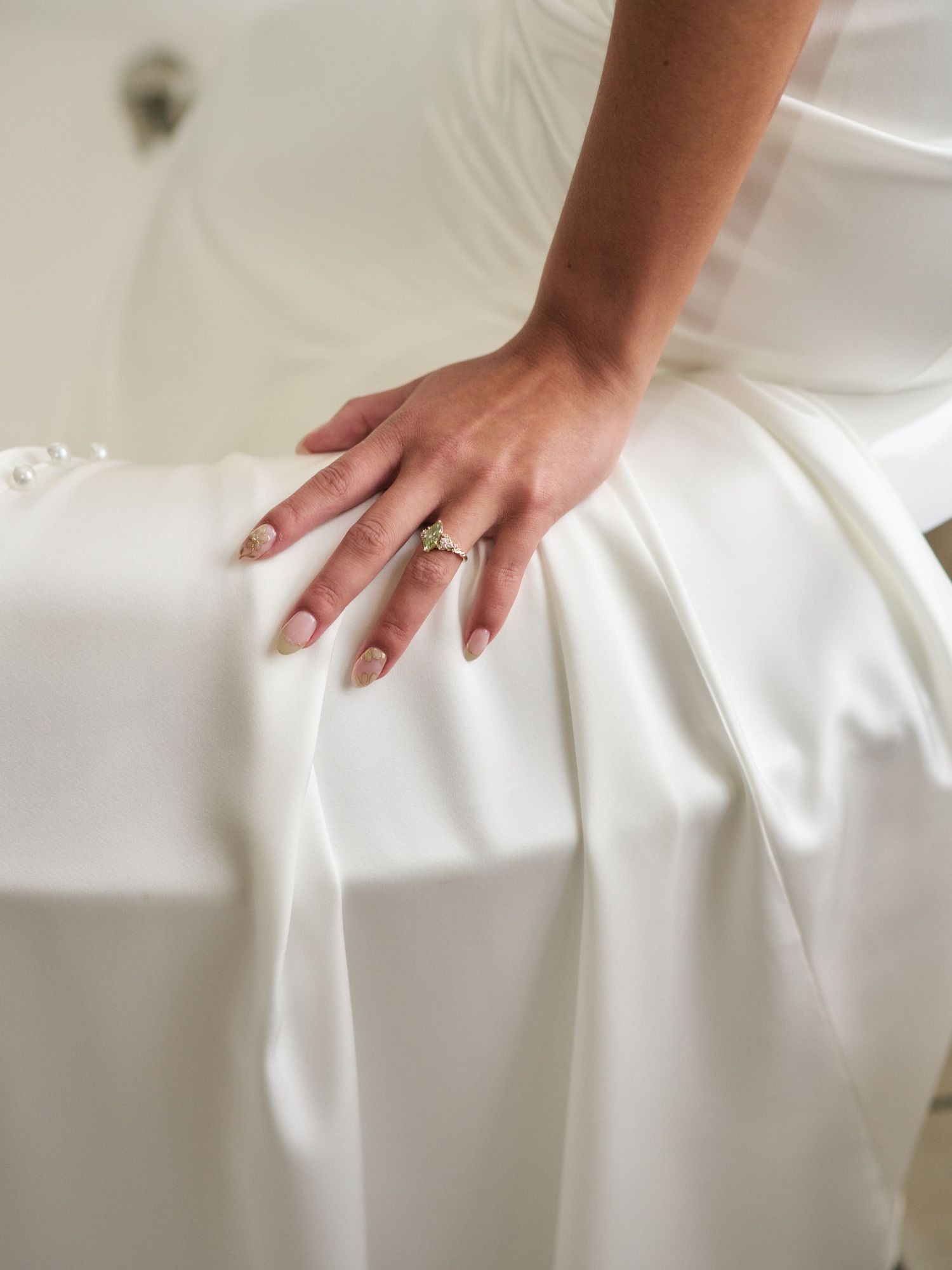 Close-up of a bride's hand with an elegant engagement ring resting on a white satin wedding dress.