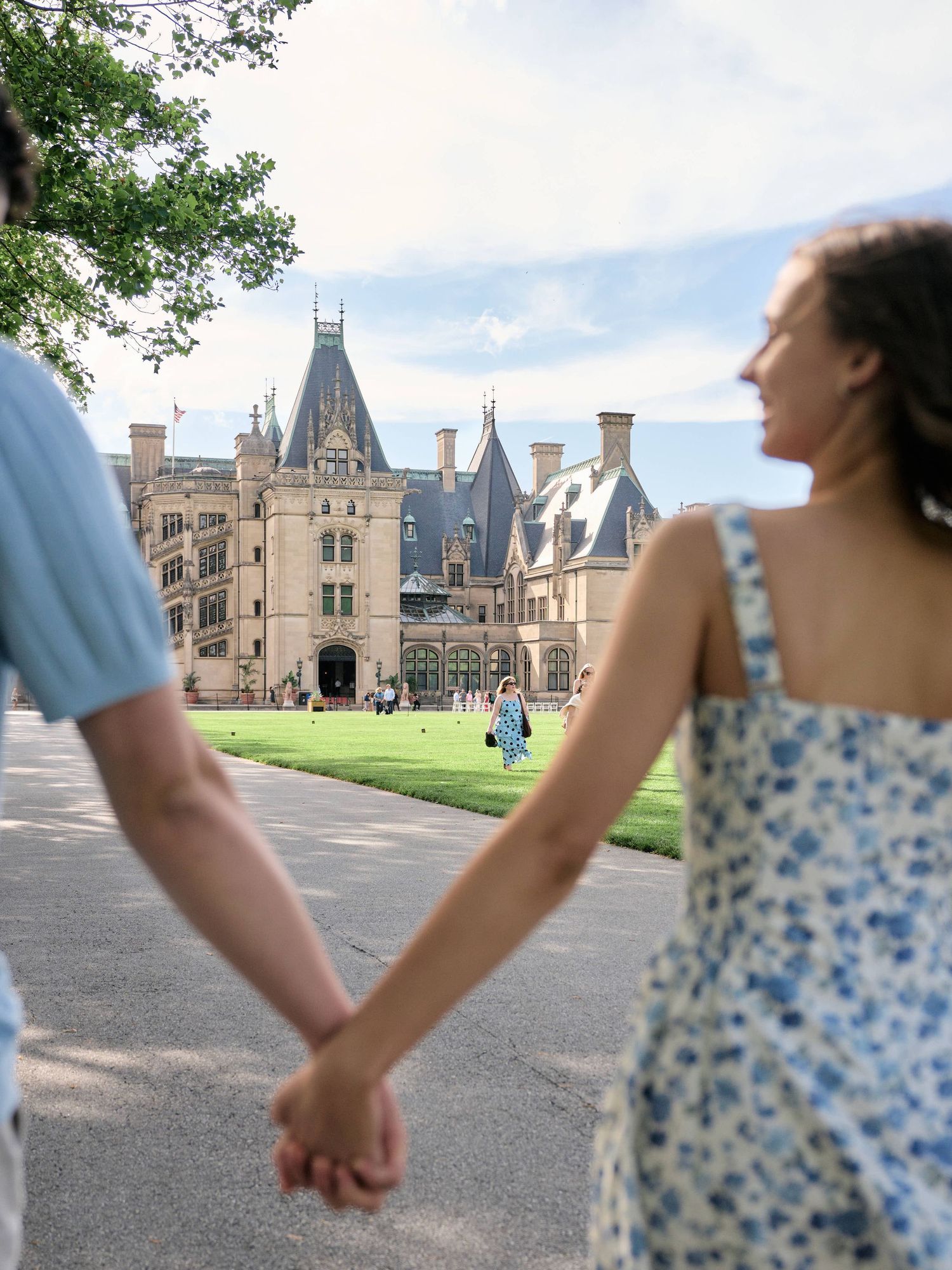 Couple holding hands walking toward Biltmore Estate mansion on a sunny day in Asheville, NC.