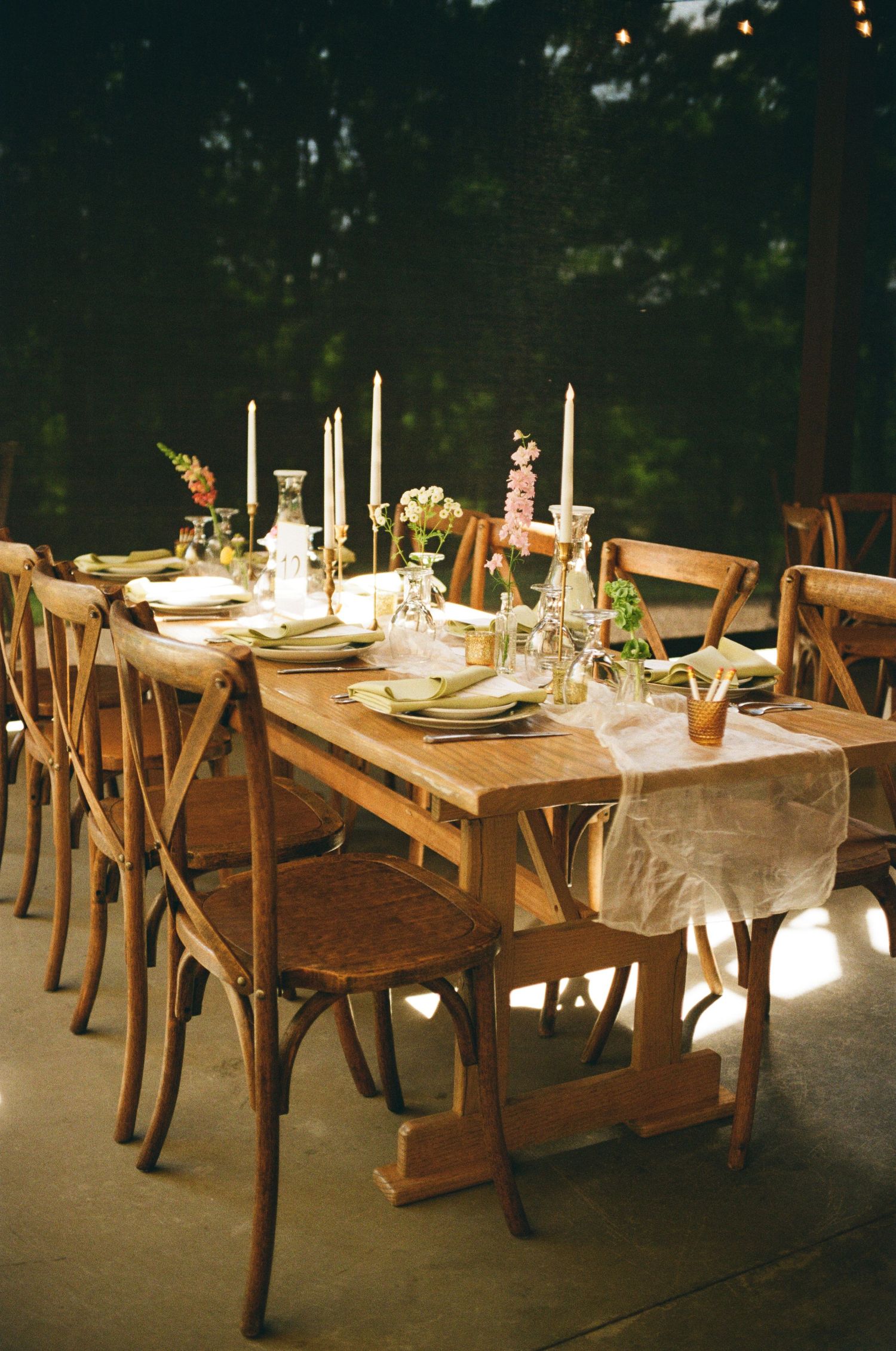 Rustic wooden dining tables set for an elegant outdoor evening dinner with white tablecloths and glowing candlelight.