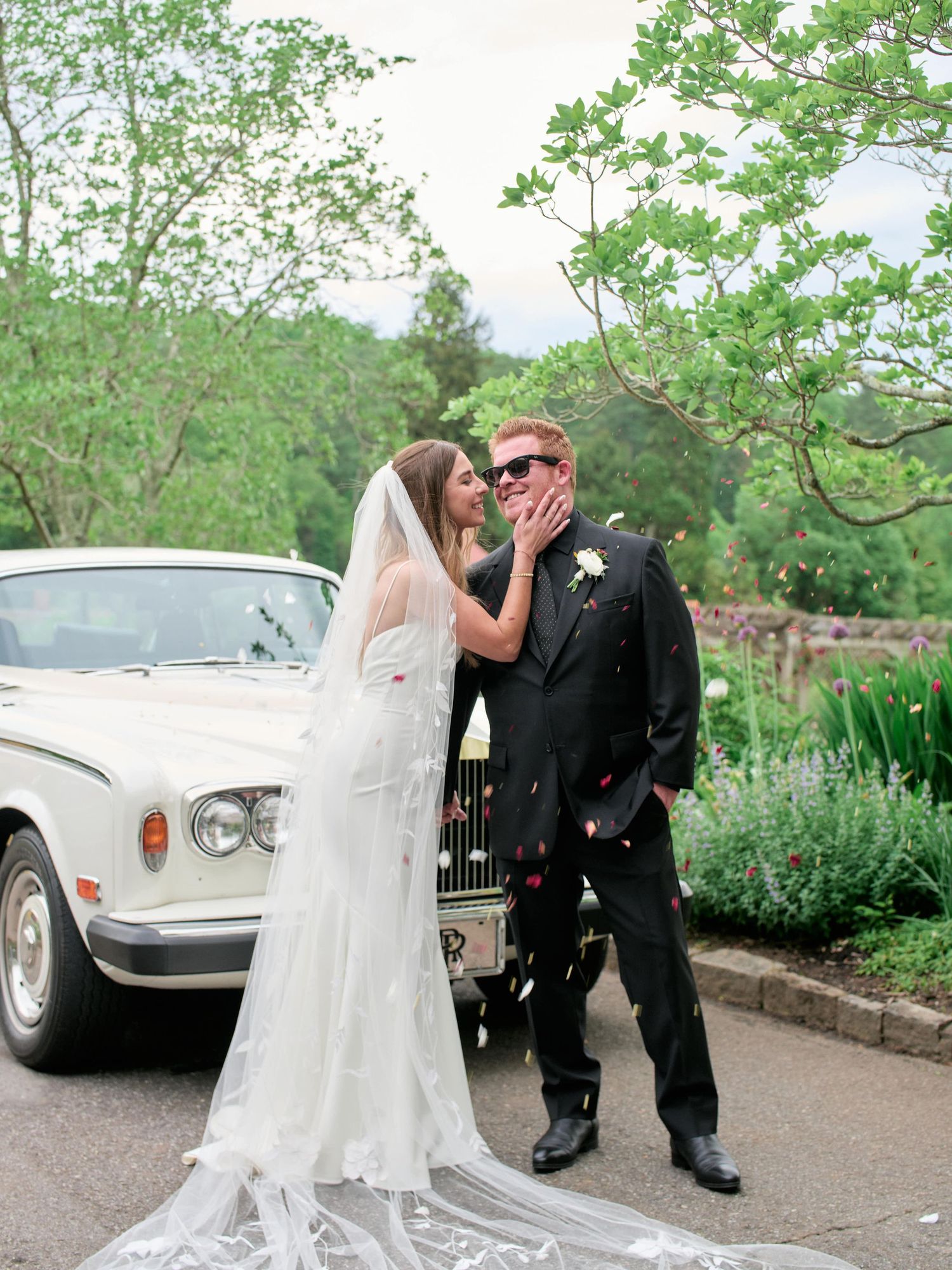 A romantic wedding couple shares intimate moments in front of a classic vintage car in a lush garden setting.