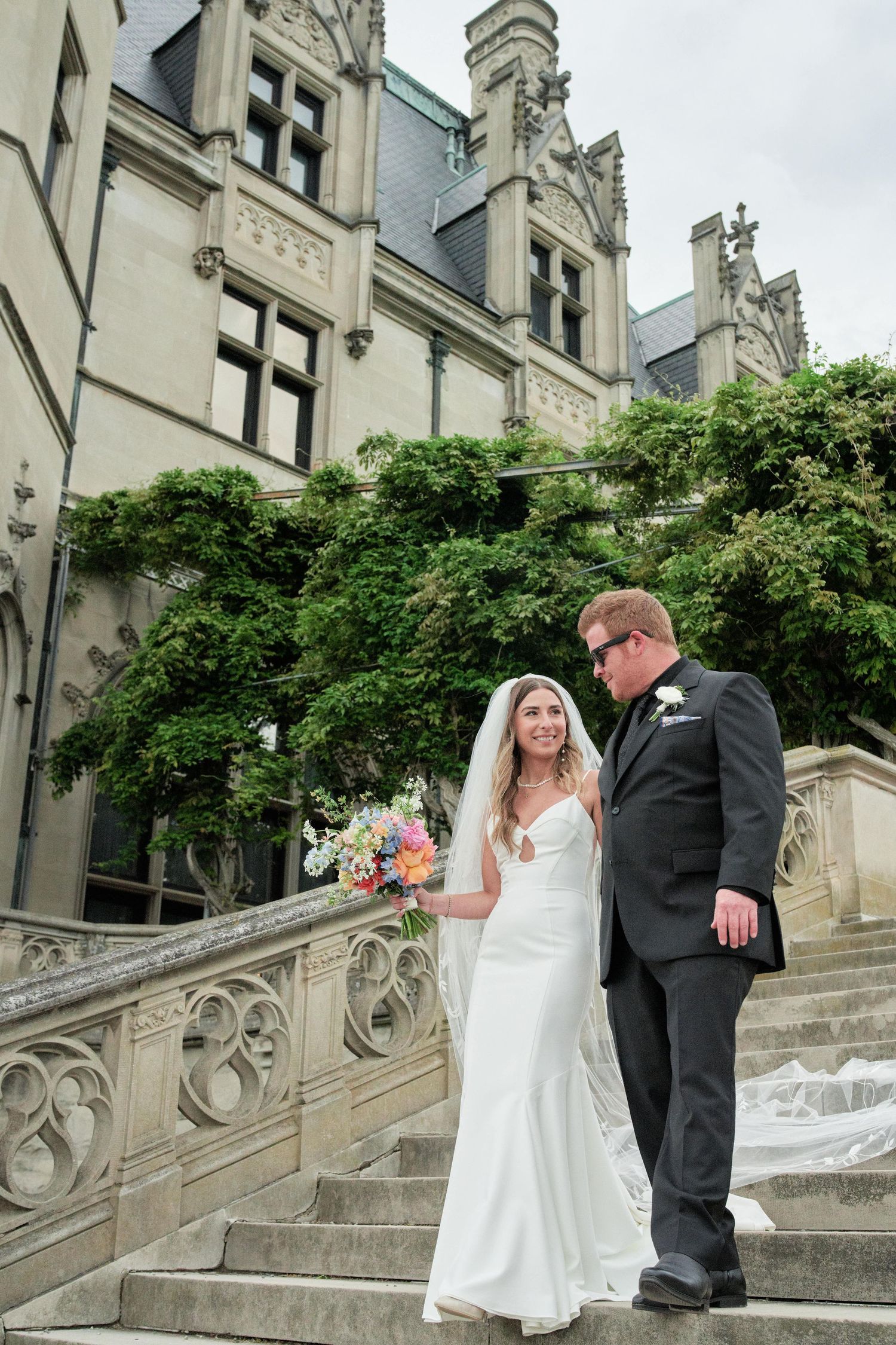 Bride and groom share romantic moments on ornate stone steps of historic building with ivy-covered walls.