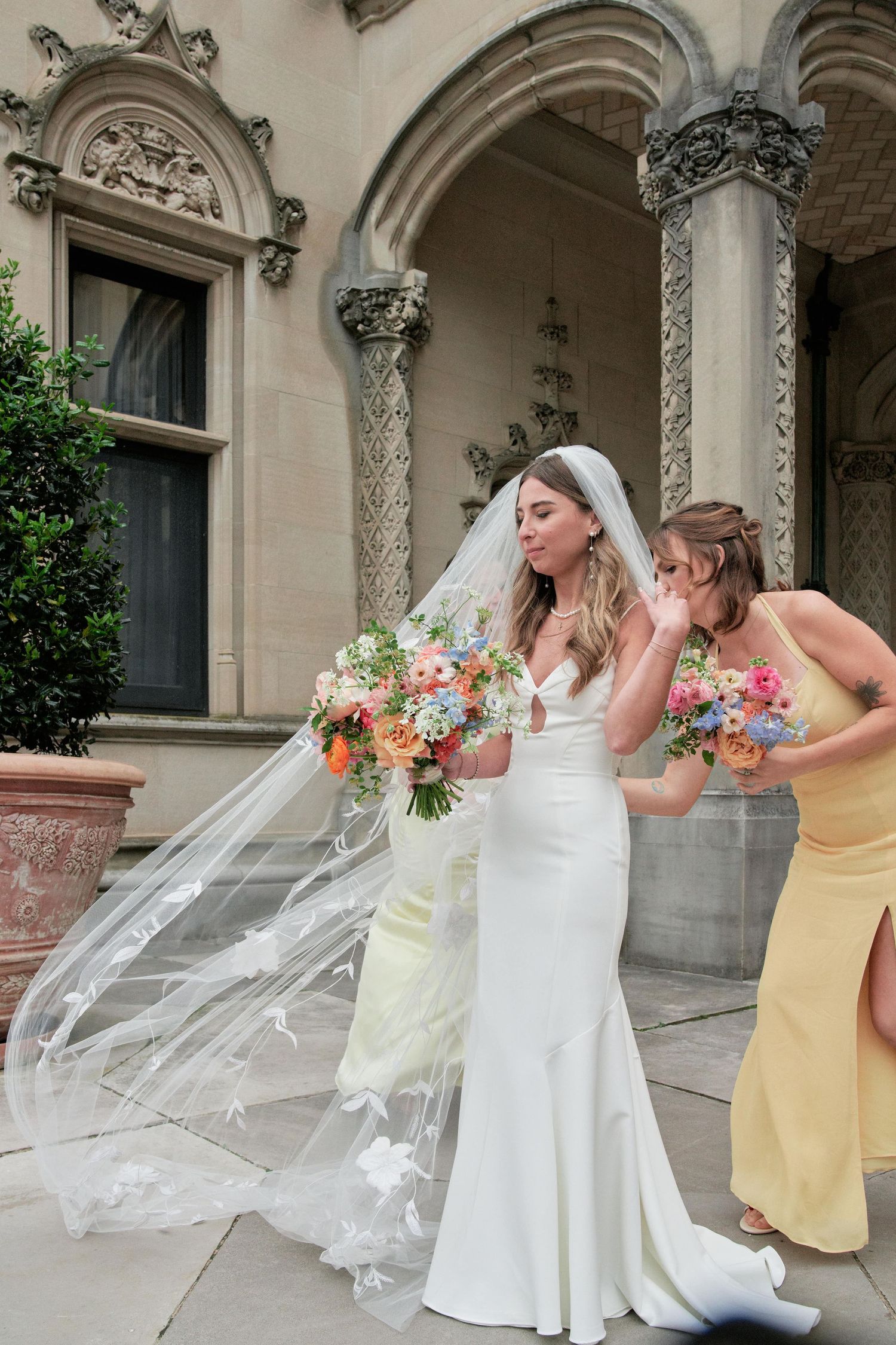 Wedding party stands on ornate stone portico with flowing veil and colorful floral bouquets in elegant outdoor setting.