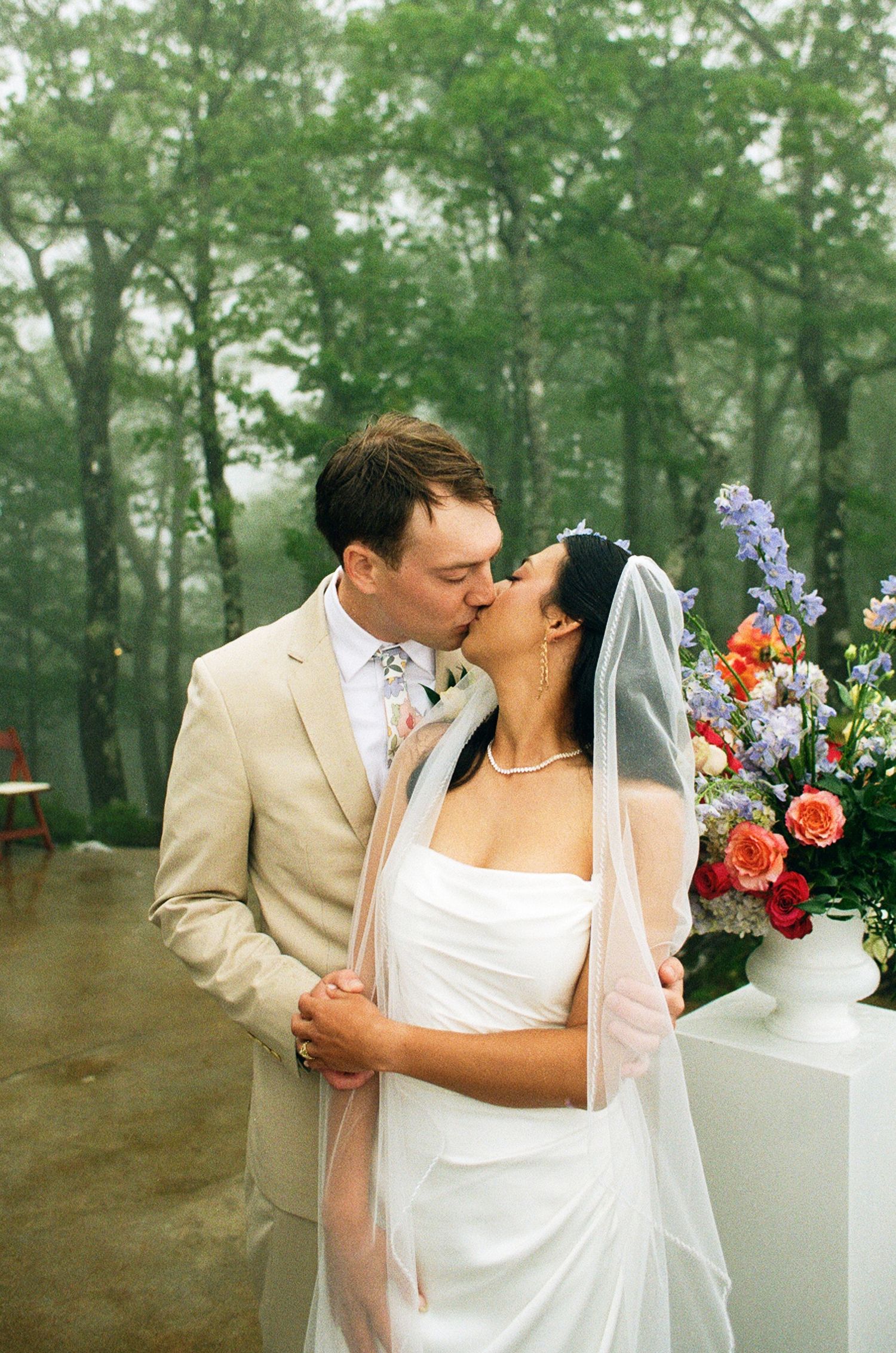 A bride and groom share a romantic kiss outdoors on their wedding day, surrounded by misty trees and colorful floral arrangements.