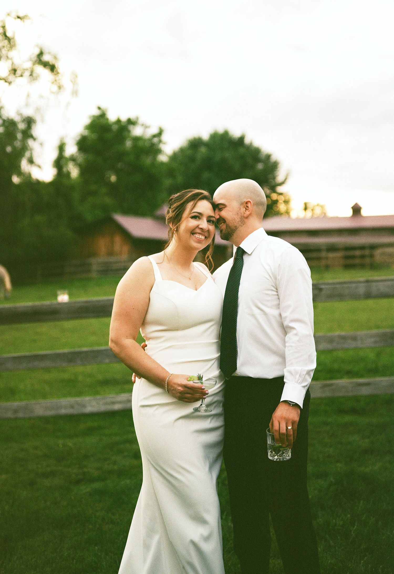 Bride in white gown and groom in white shirt with dark tie embrace outdoors at a rustic farm wedding venue.