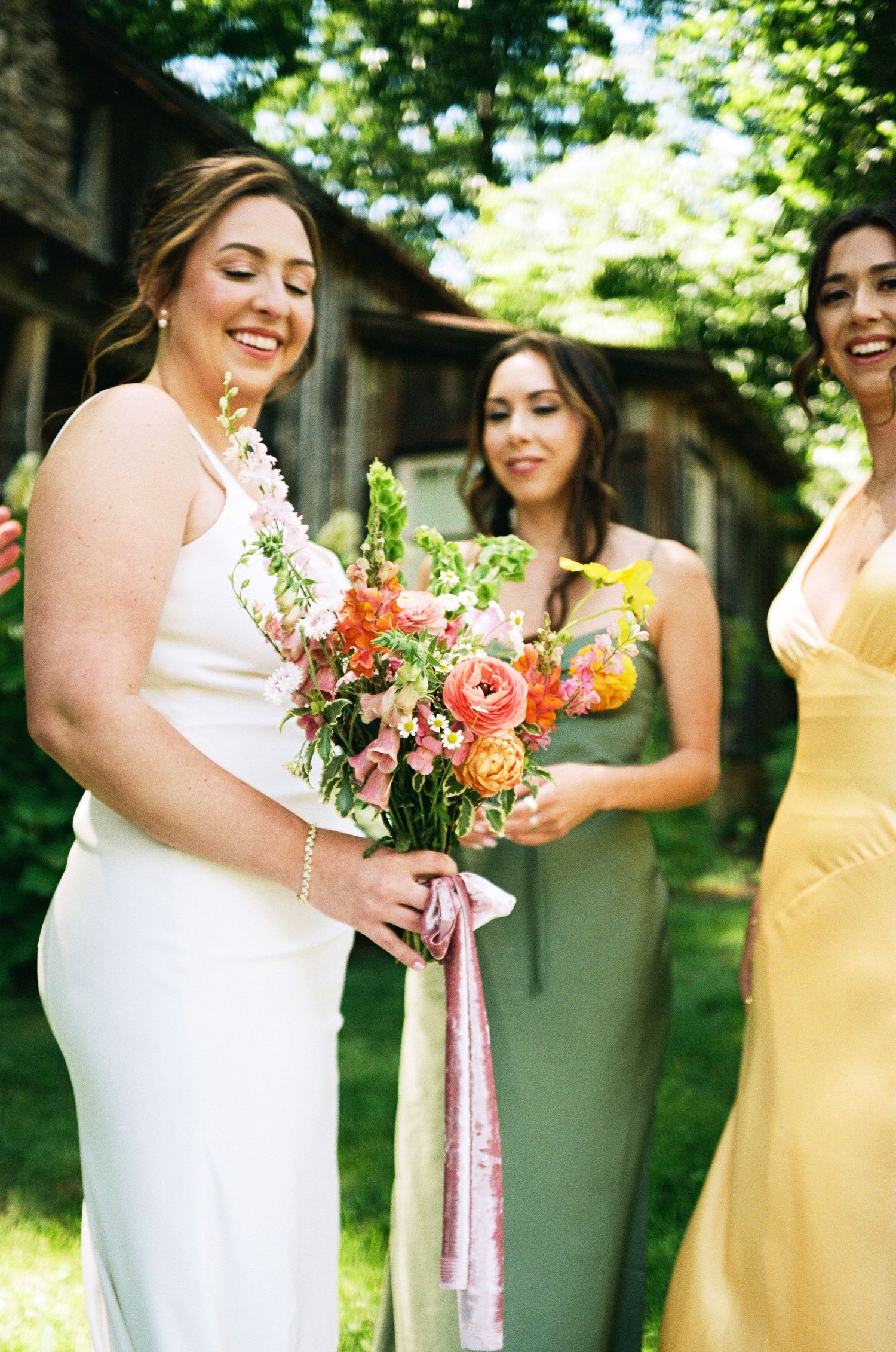 Bride in white gown holds colorful bouquet with pink ribbons, flanked by bridesmaids in sage and yellow dresses outdoors.