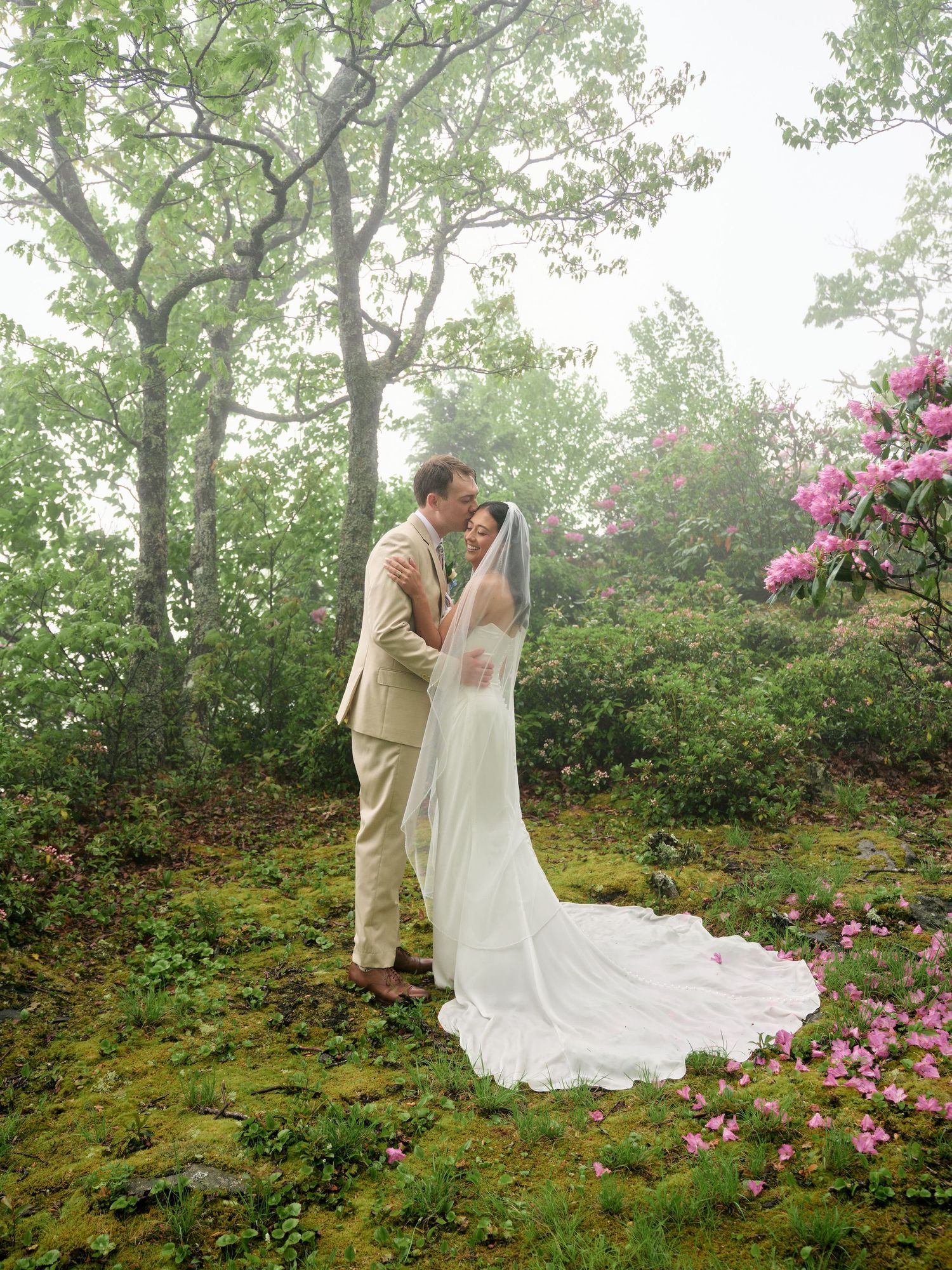 A couple shares a romantic kiss in a lush green forest setting, bride in white gown with veil, groom in tan suit.