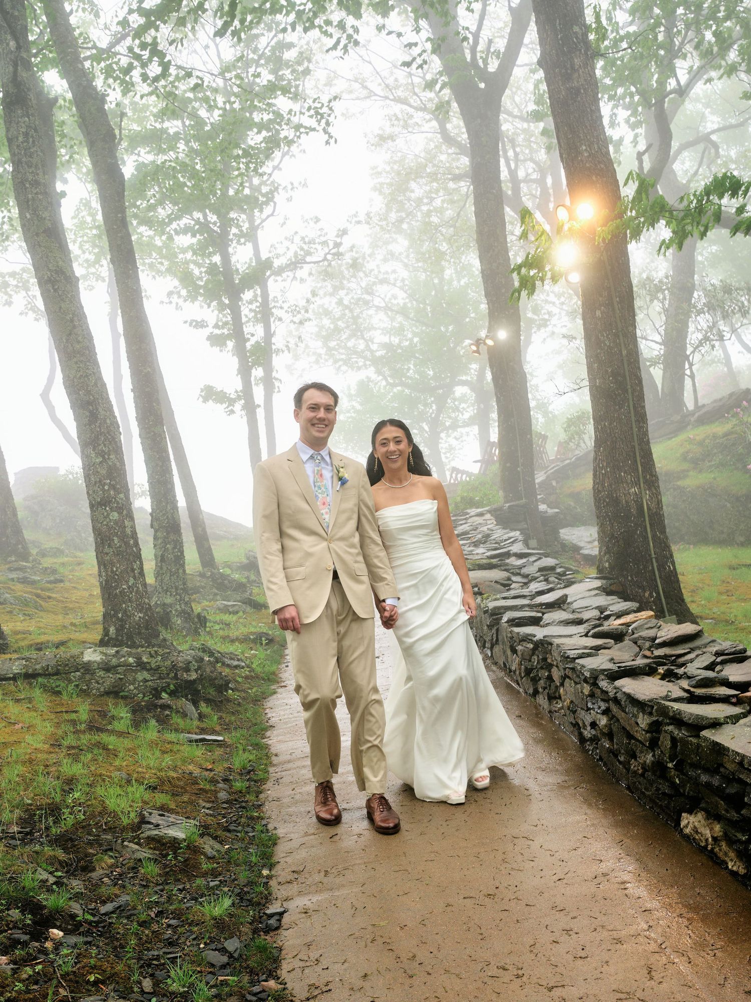 A bride and groom walk hand-in-hand on a misty forest path beside a stone wall on their wedding day.