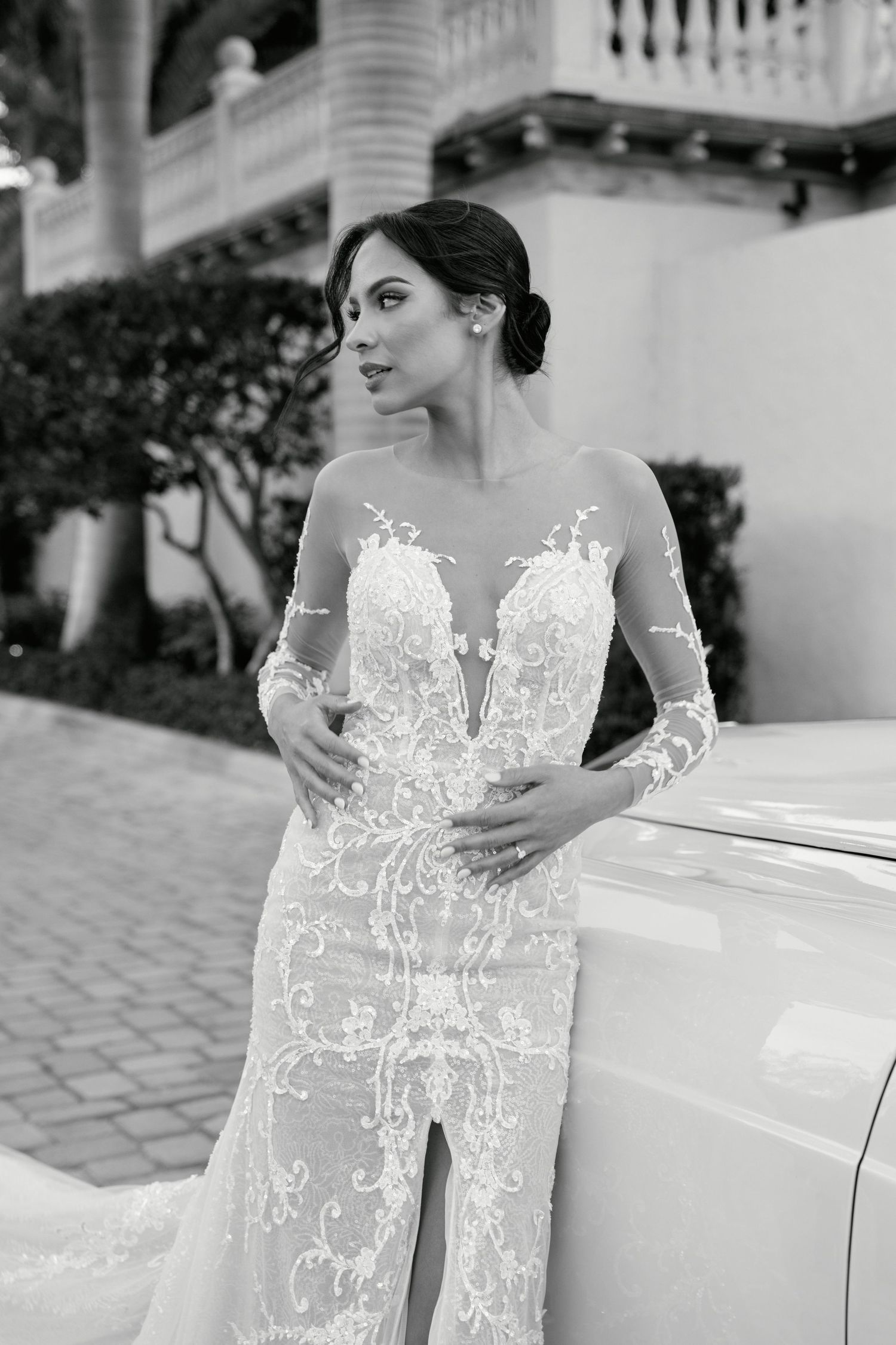 Black and white portrait of bride in fitted lace gown beside classic car.