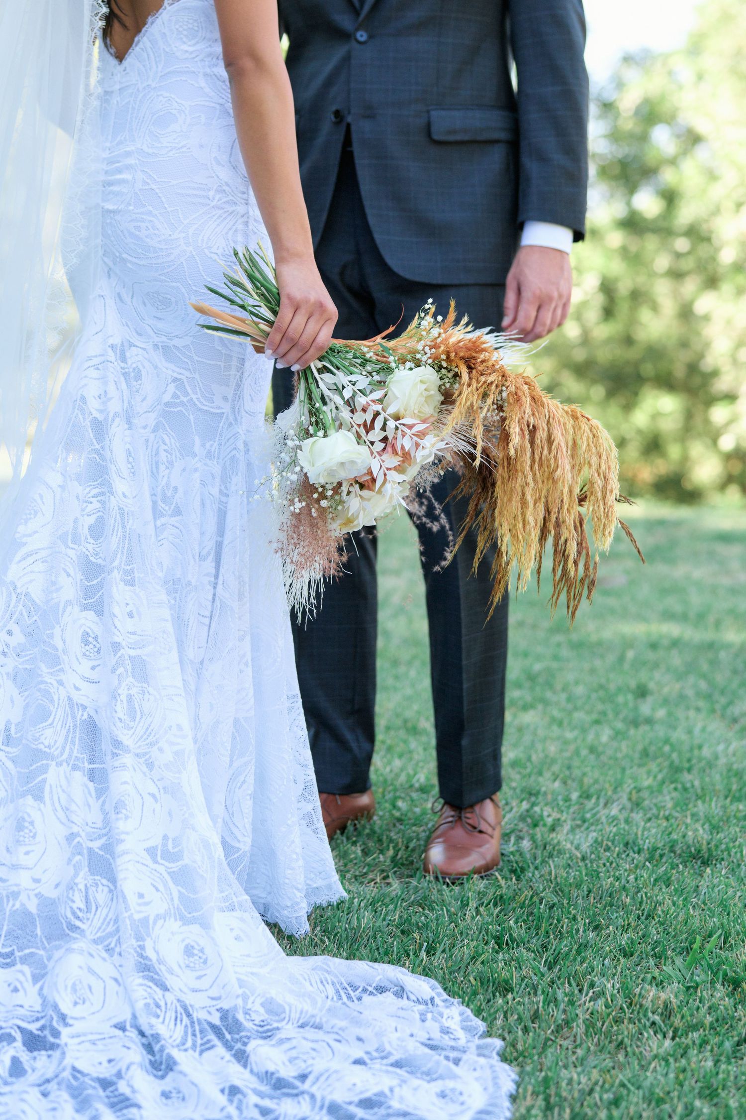 Close-up of bride's lace wedding dress and dried floral bouquet against green grass.