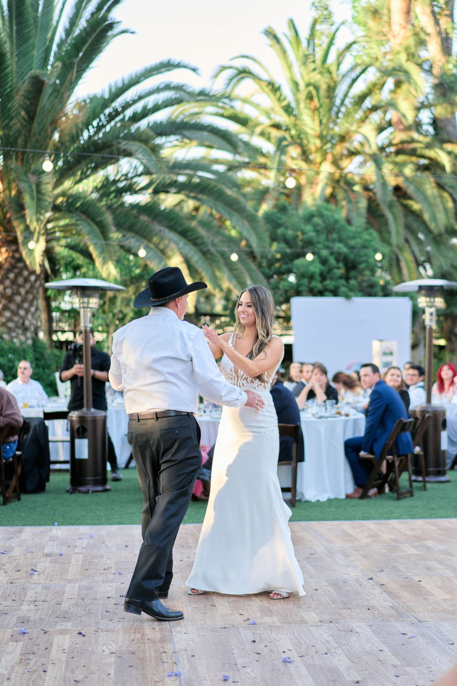 A couple dances on a wooden floor during an outdoor wedding reception surrounded by palm trees and white tables.
