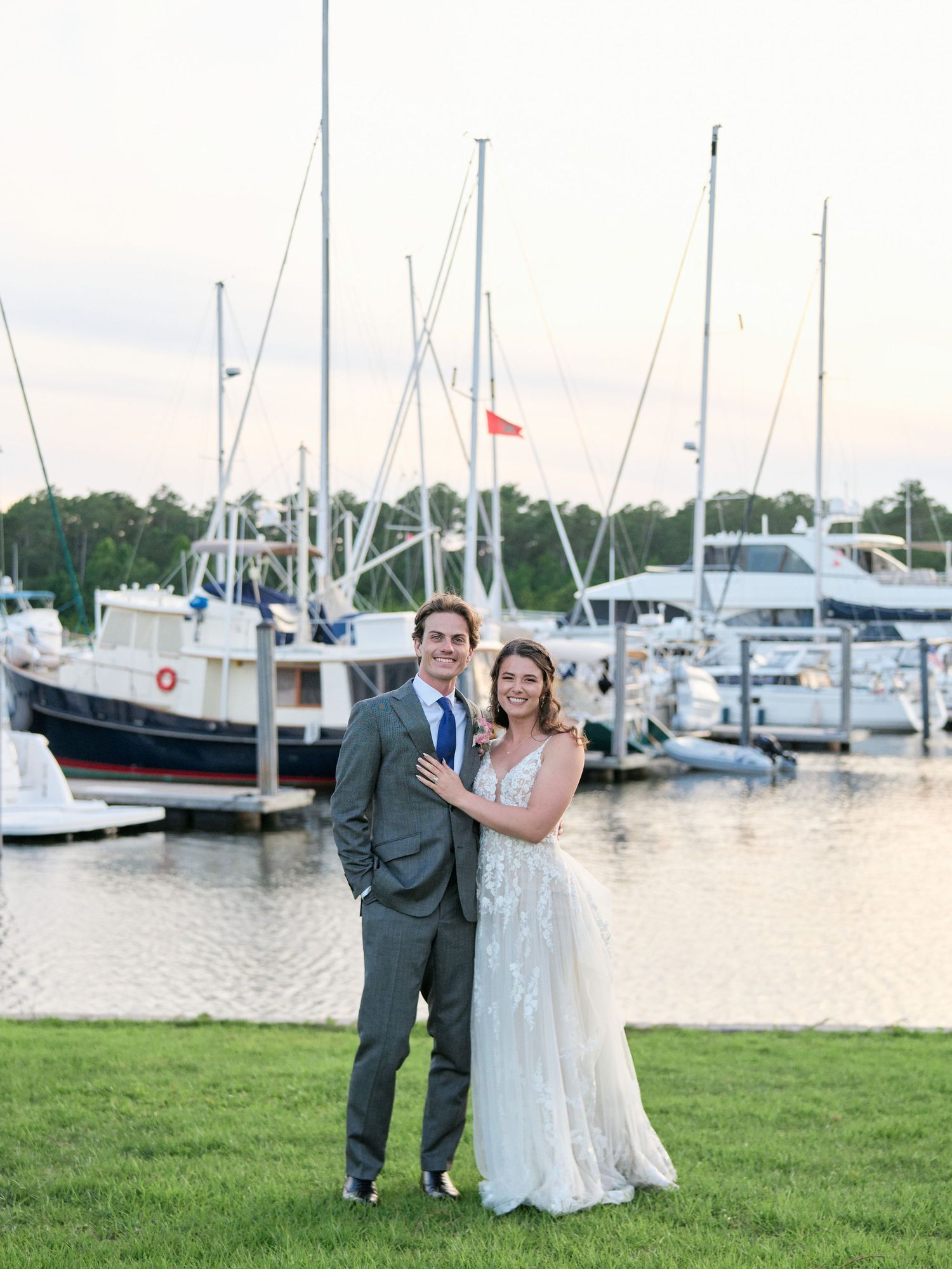 A couple shares a romantic moment on grass near a marina with boats and yachts docked in the background at sunset.