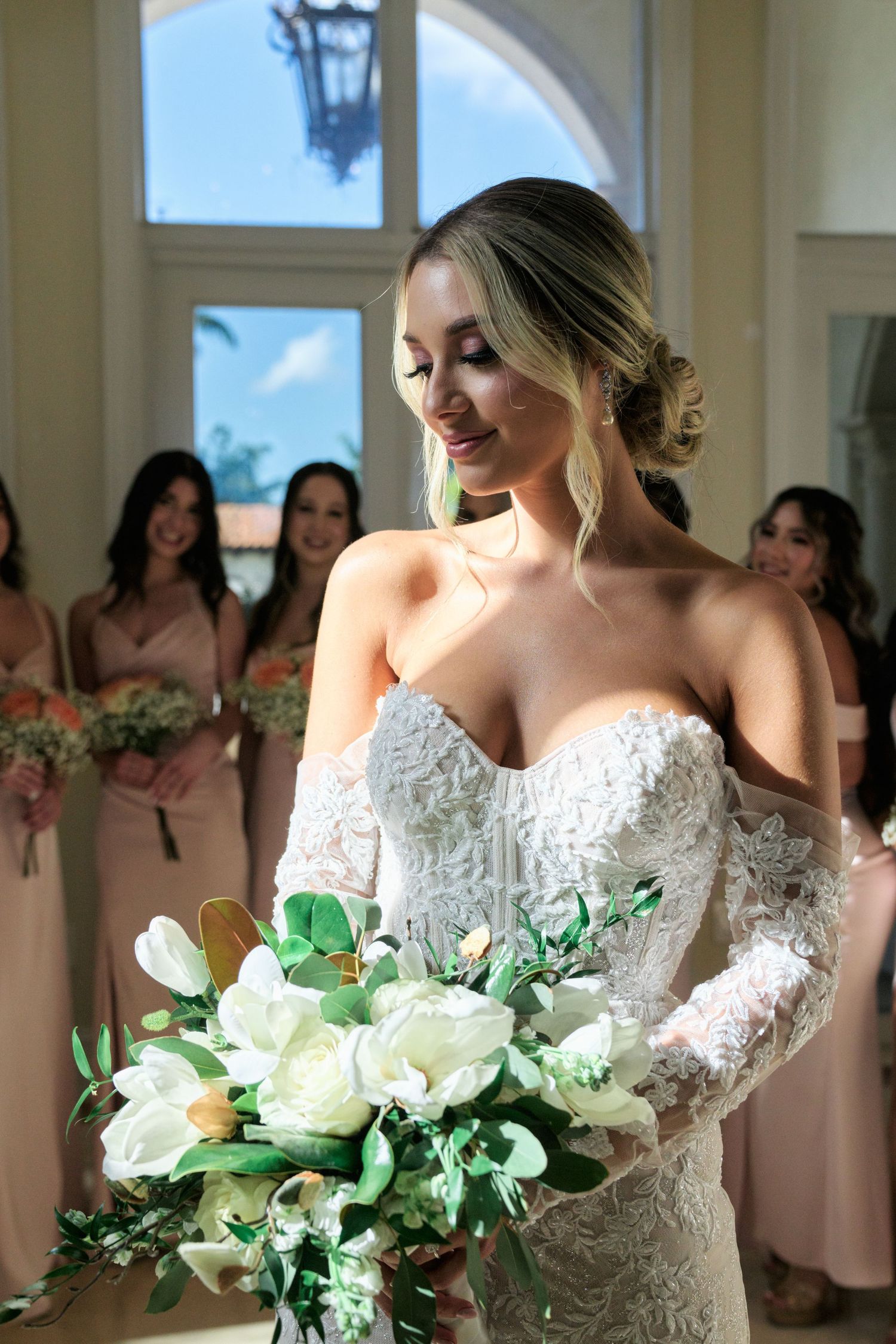 A bride wearing a white lace dress holds a bouquet of white roses and greenery.