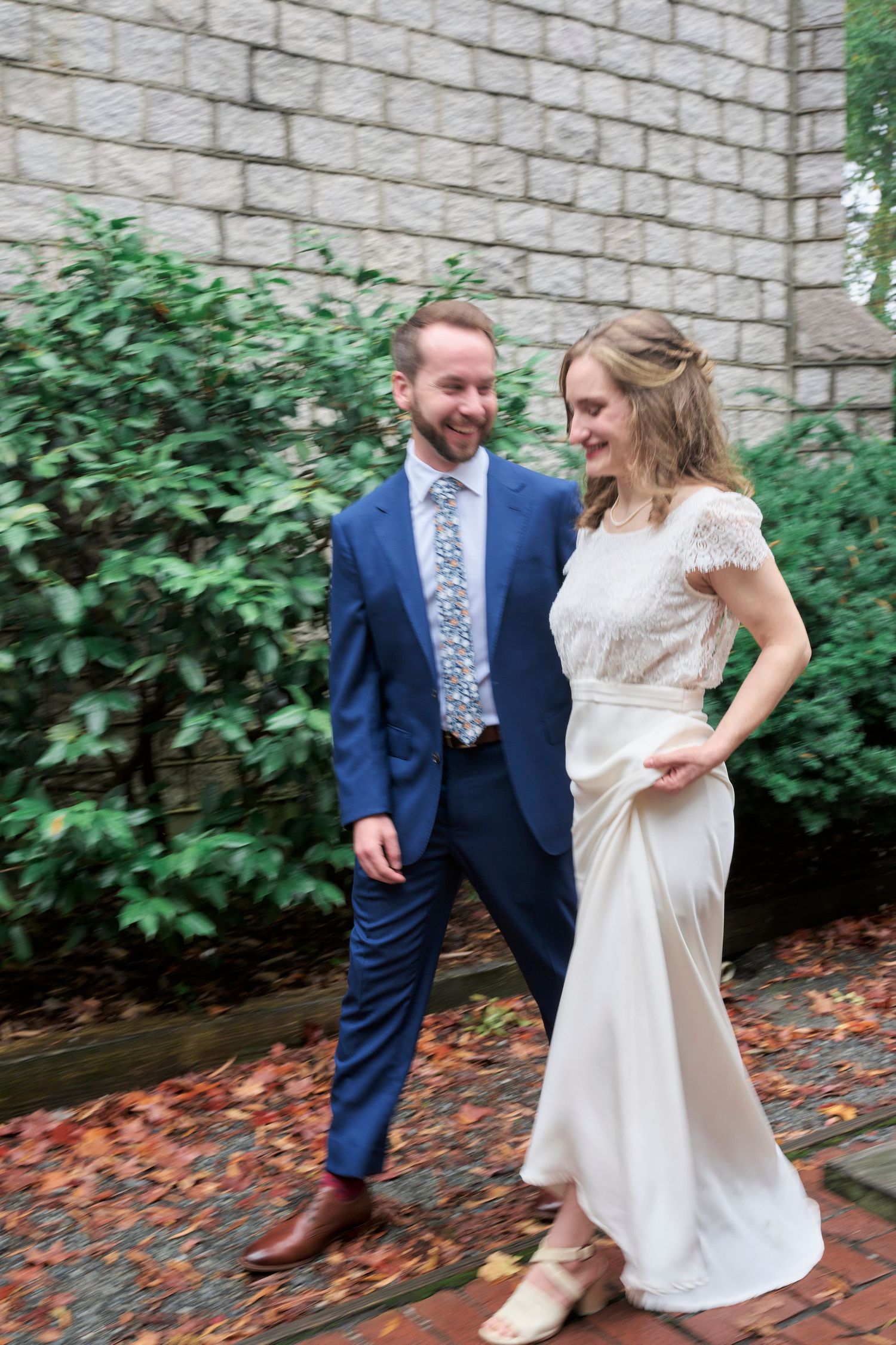 A couple in wedding attire share a joyful moment together on a brick walkway near stone walls and greenery.