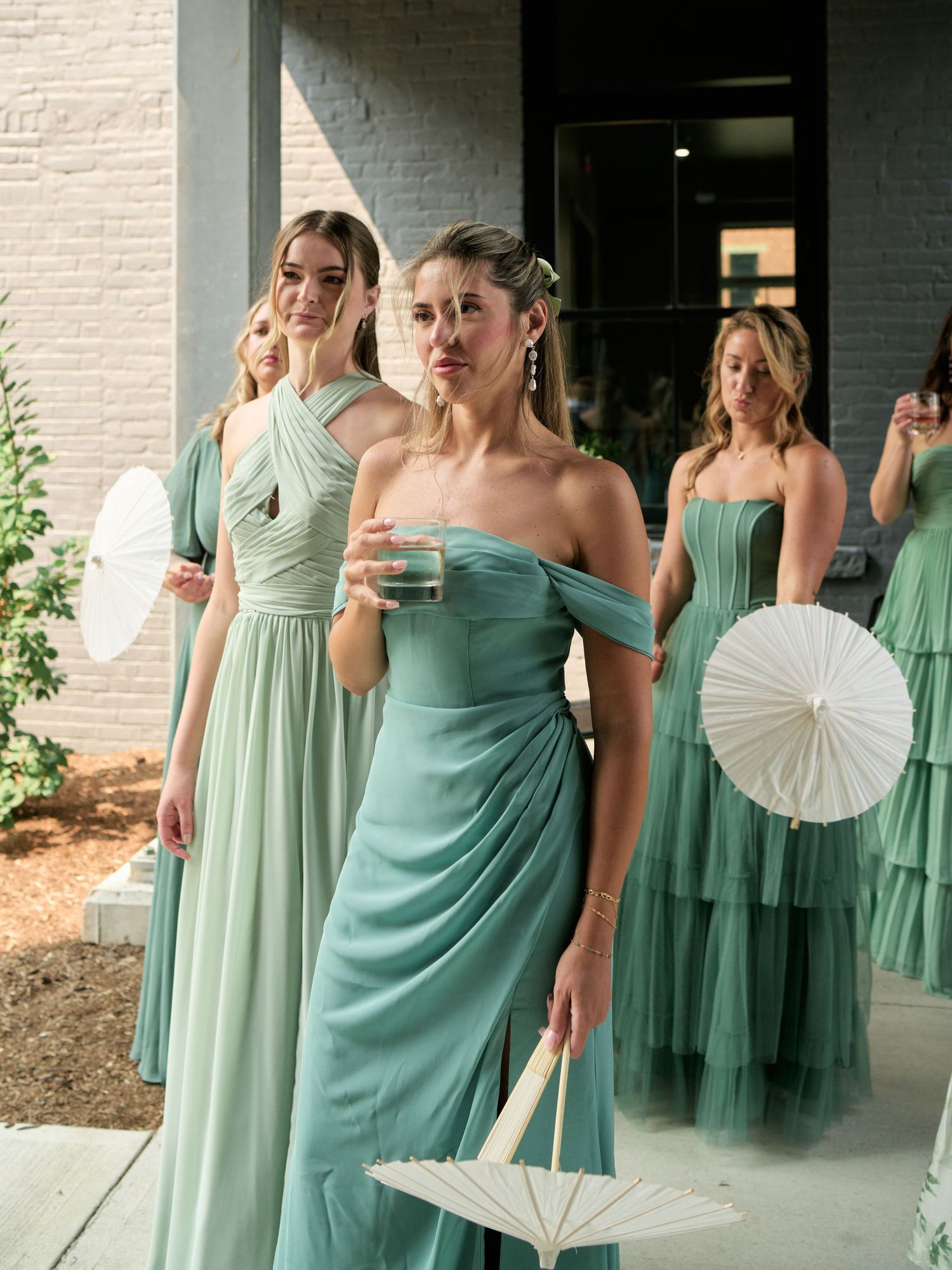 Elegant bridesmaids in seafoam green dresses hold white paper parasols while posing outside a brick building.