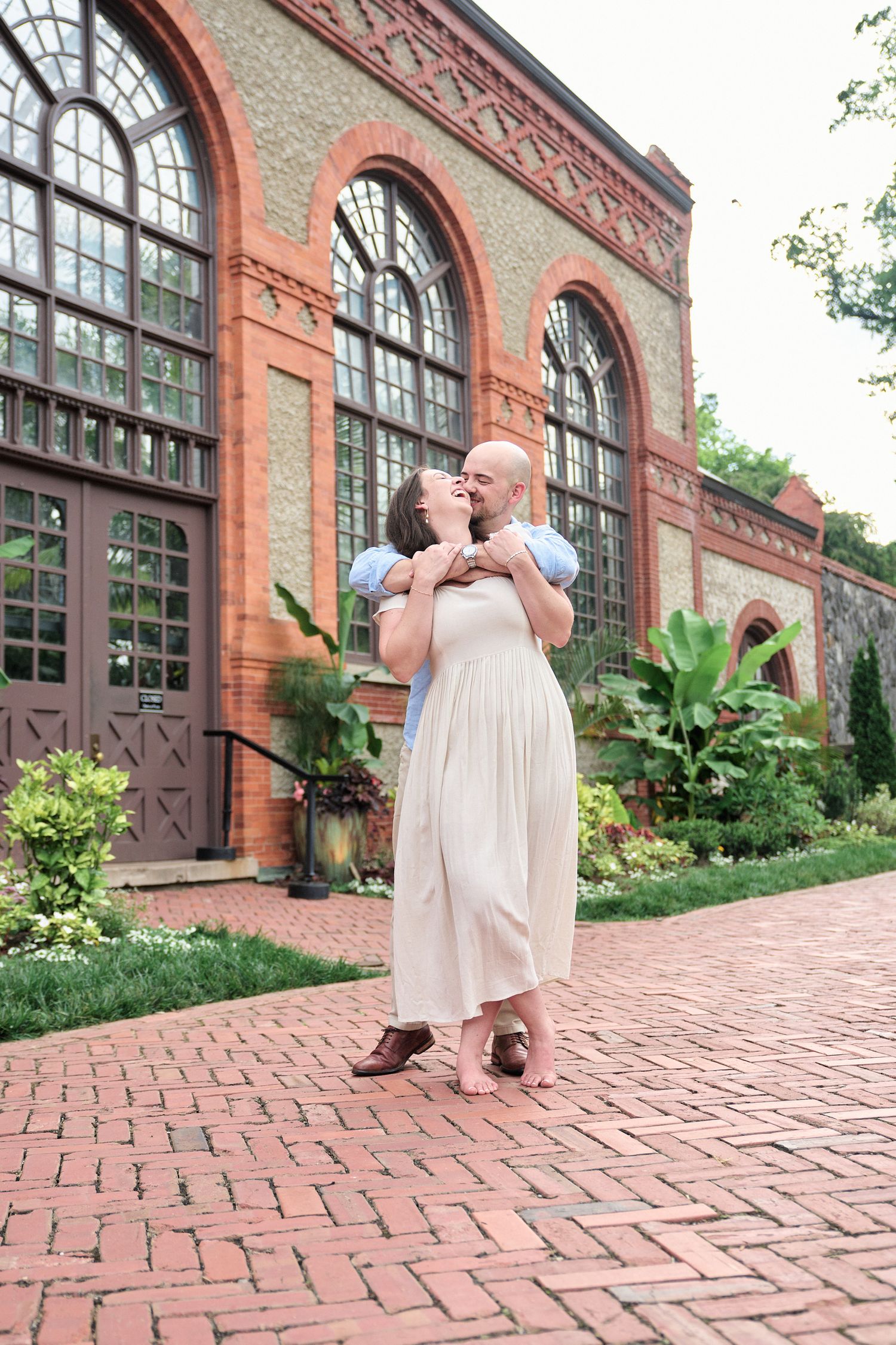 A romantic moment captured in front of an ornate brick building with arched windows and tropical plants.