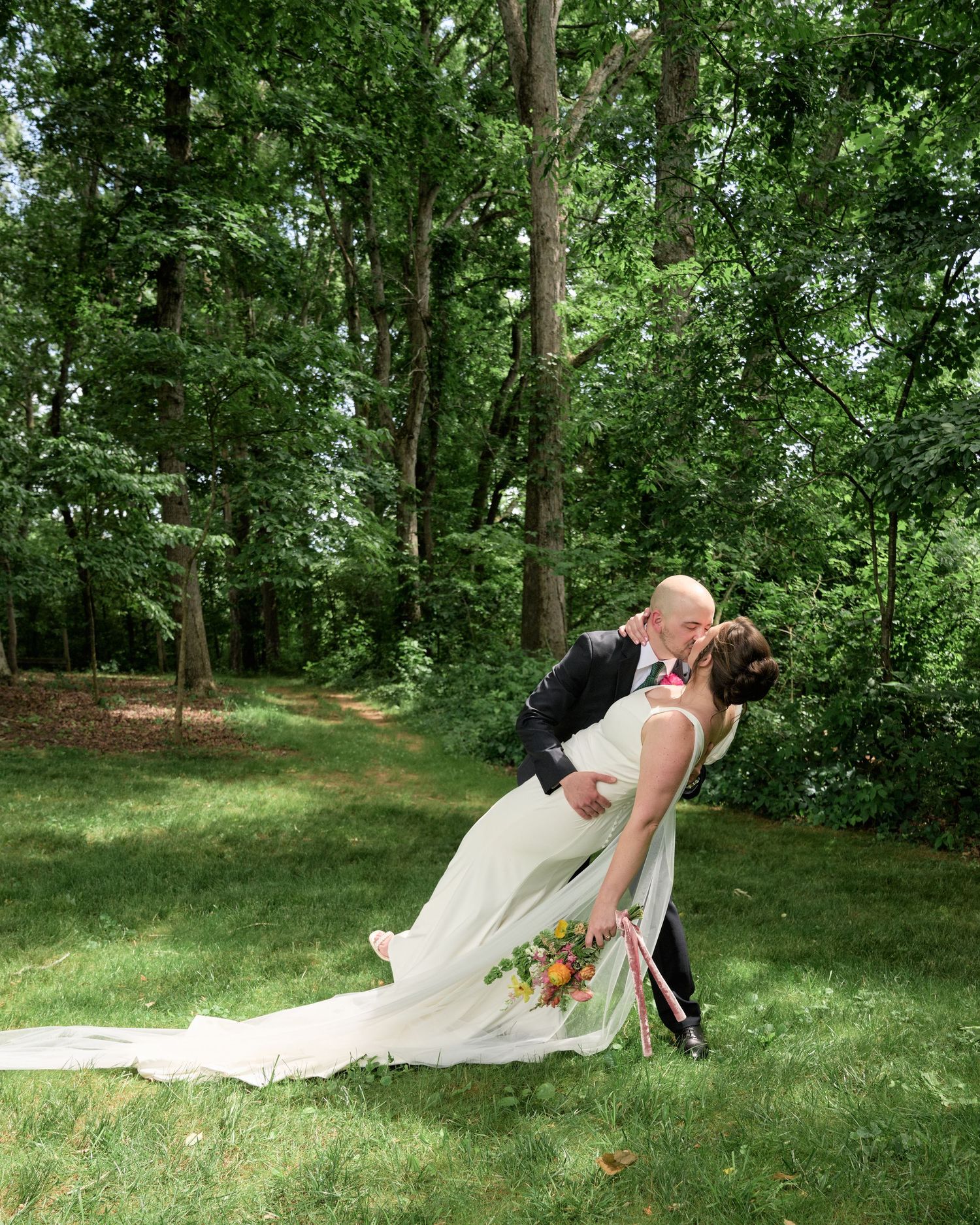 A romantic dip pose between newlyweds in a forest setting with the bride's dress trailing on green grass.