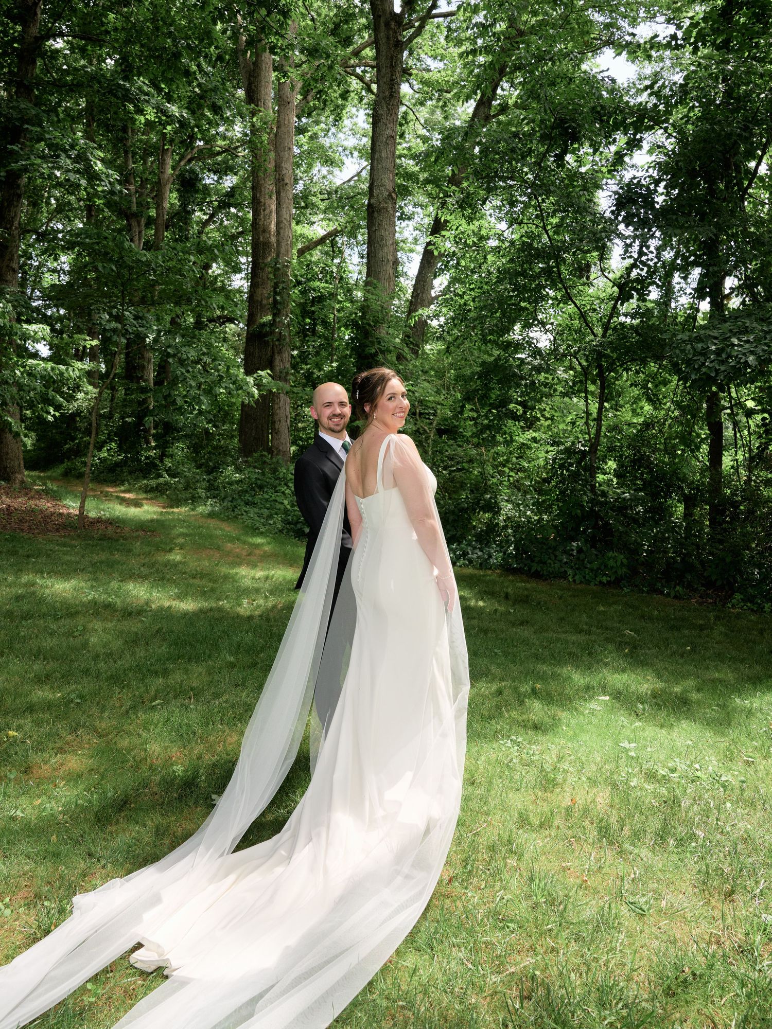 Couple stands together in elegant wedding attire with long flowing veil against backdrop of trees.