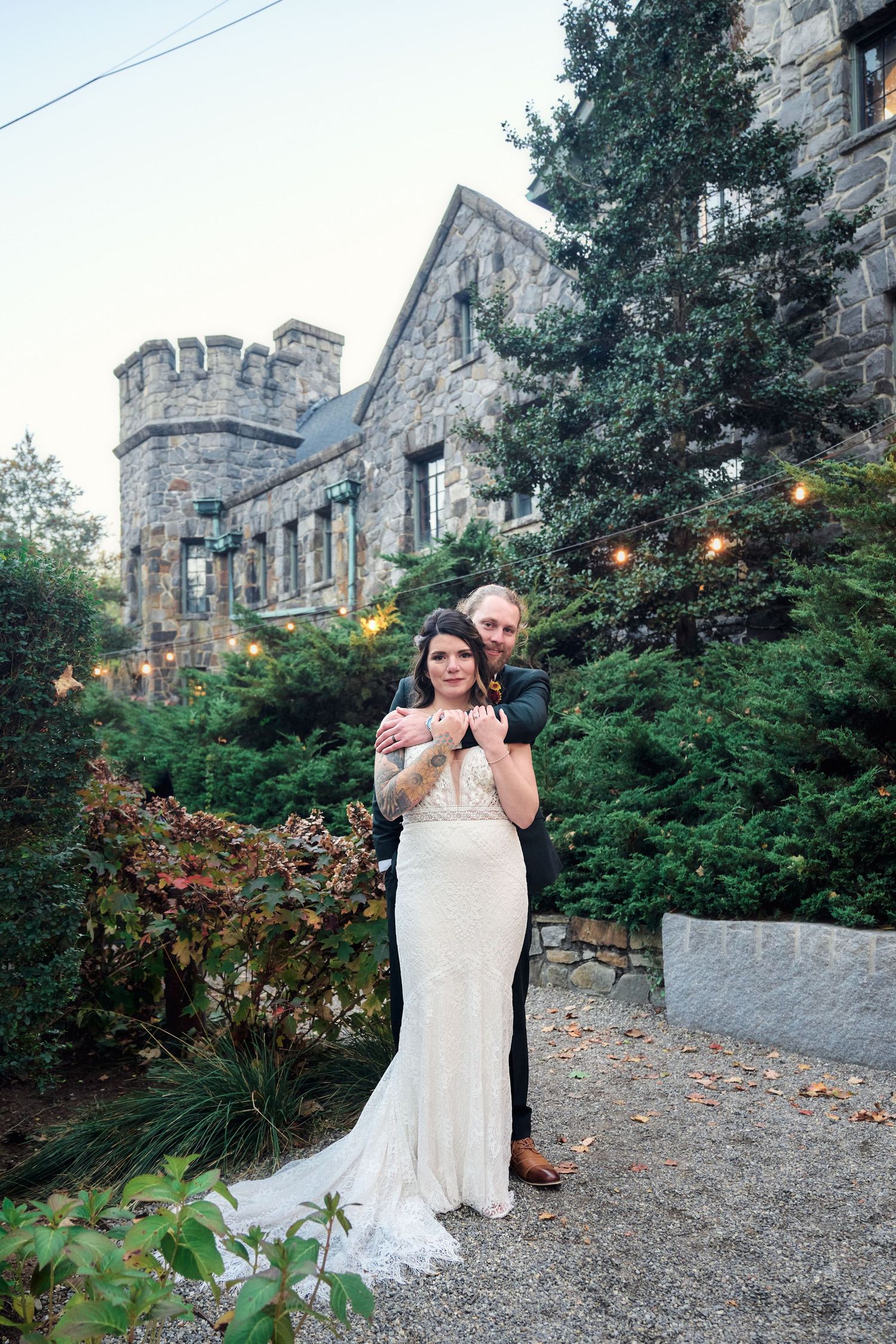 A romantic wedding photo in front of a stone castle with decorative lighting and greenery.