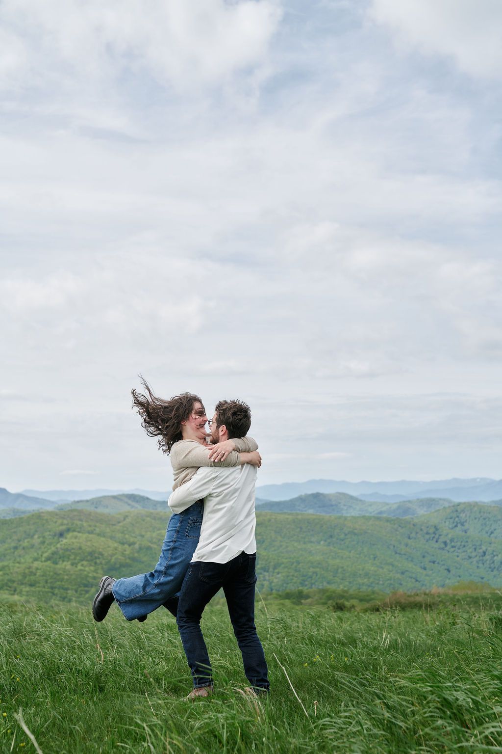 A couple dances and spins playfully in a green meadow with rolling mountains in the background.
