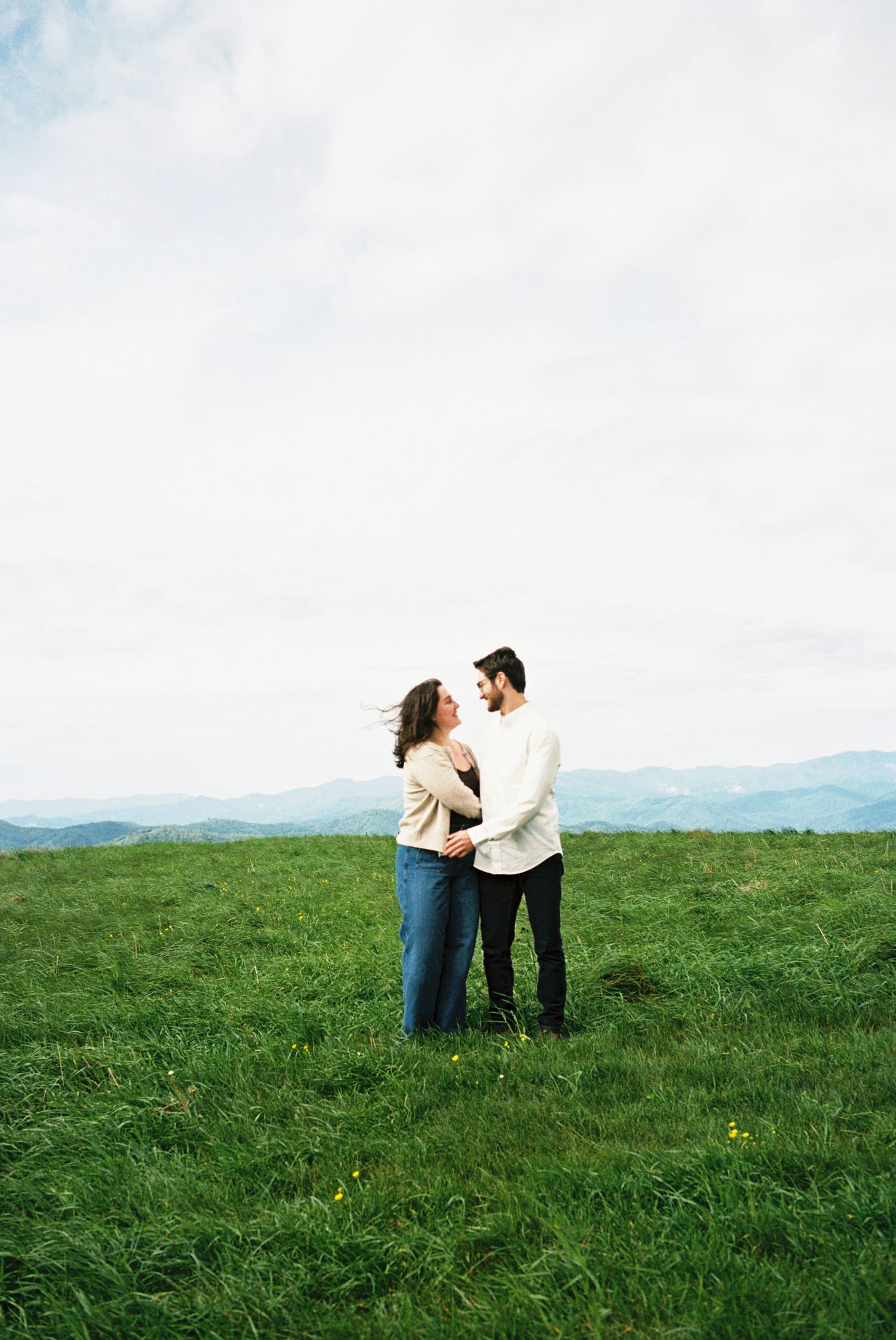 A couple embraces on a hilltop meadow against a backdrop of distant mountains and blue sky.