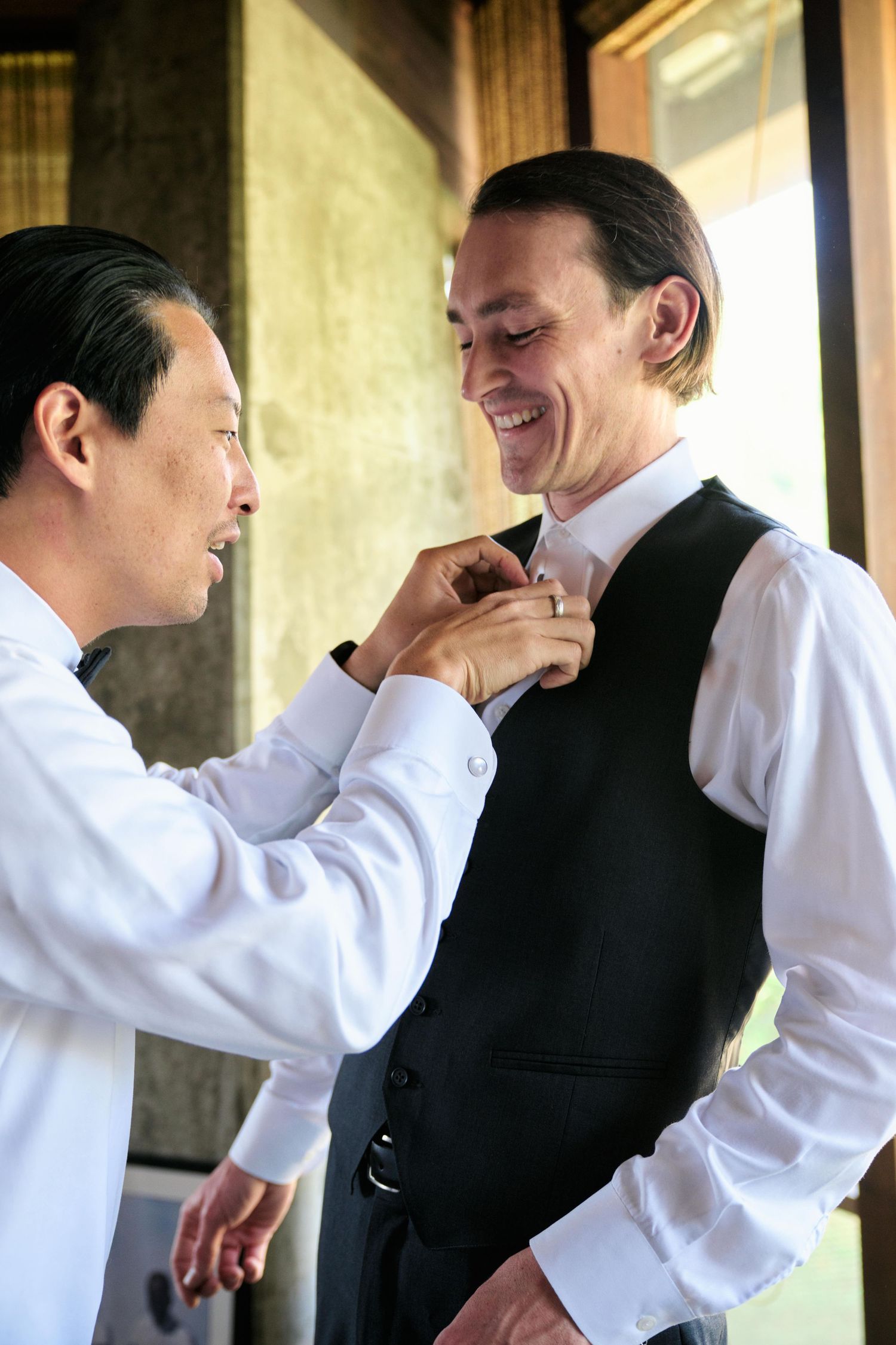 Groomsman helping adjust fellow wedding party member's bowtie while both smile.