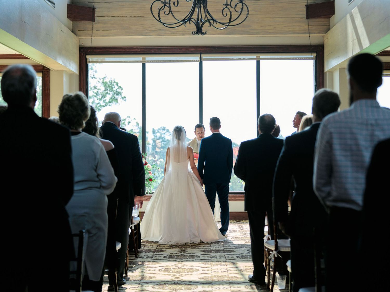 Silhouetted guests watch wedding ceremony in venue with dramatic window backdrop.