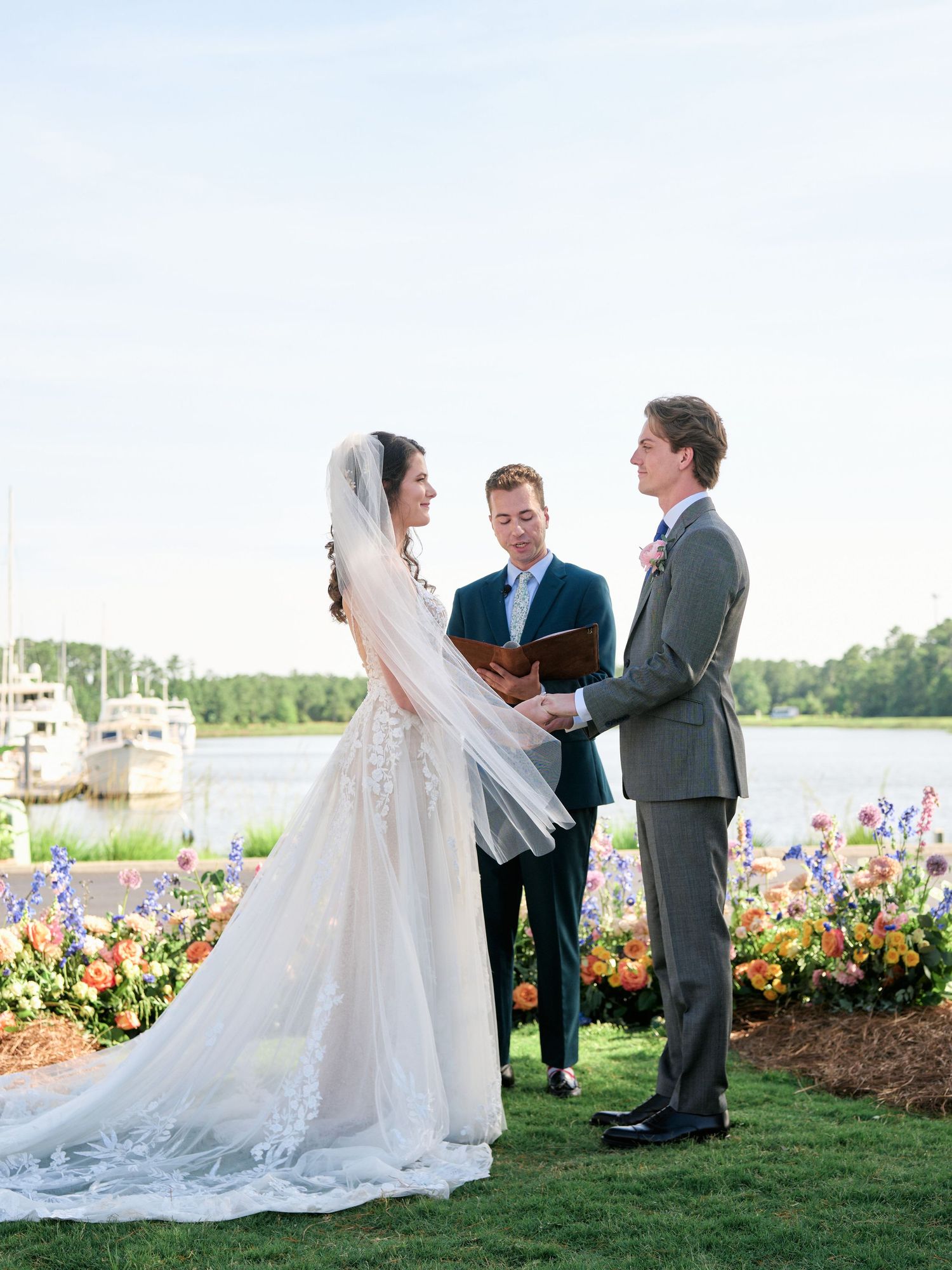 Outdoor waterfront wedding ceremony with colorful floral arrangements and a beautiful long-train lace dress against a marina backdrop.