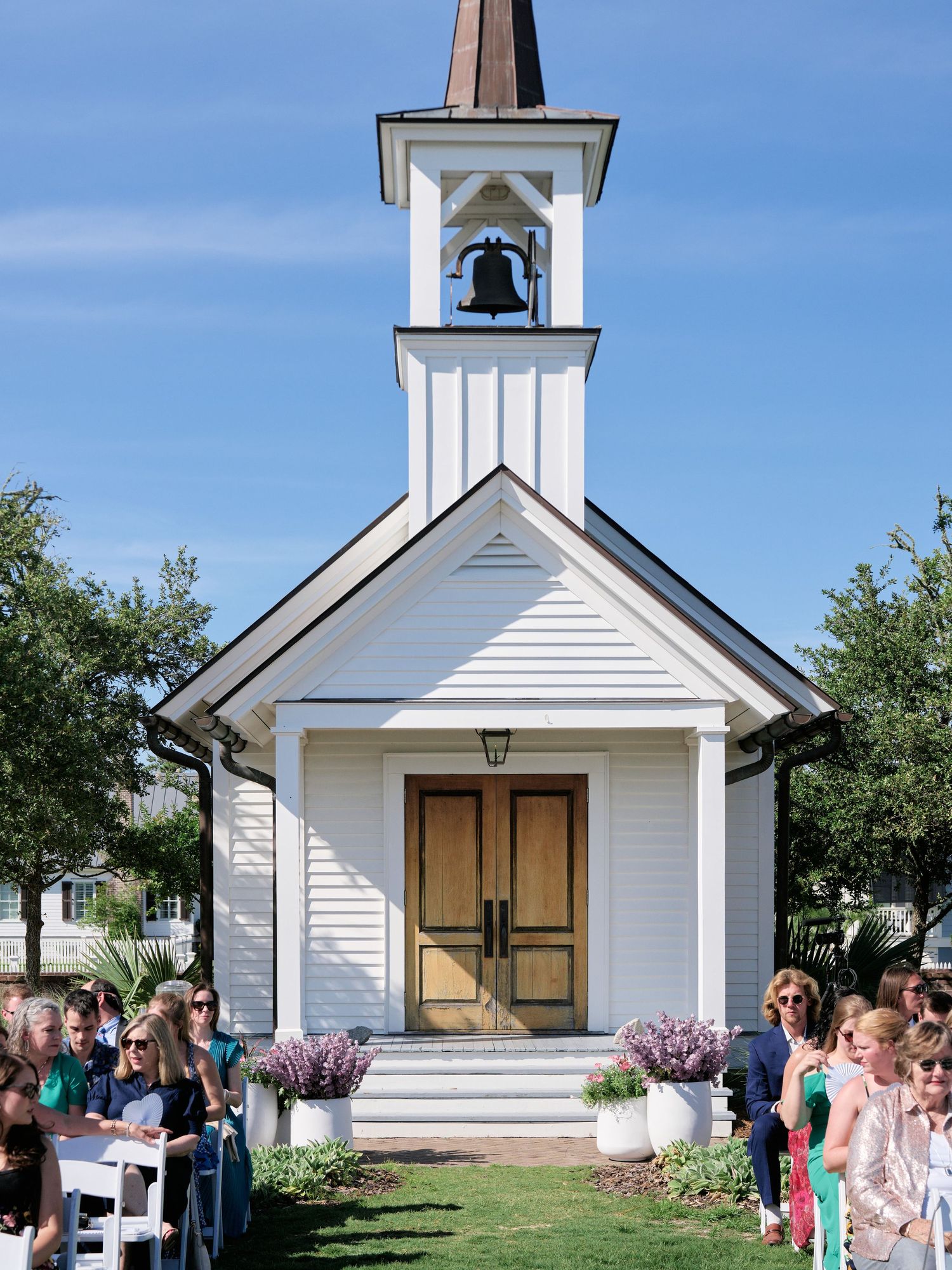 A charming white chapel with a bell tower stands at the end of an outdoor aisle lined with wedding guests under blue skies.
