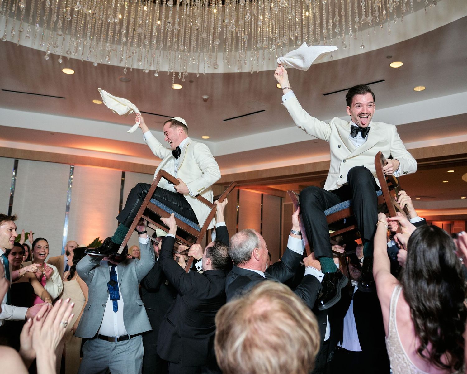 Guests celebrate at a wedding reception by lifting two people in white suits onto their shoulders while dancing.