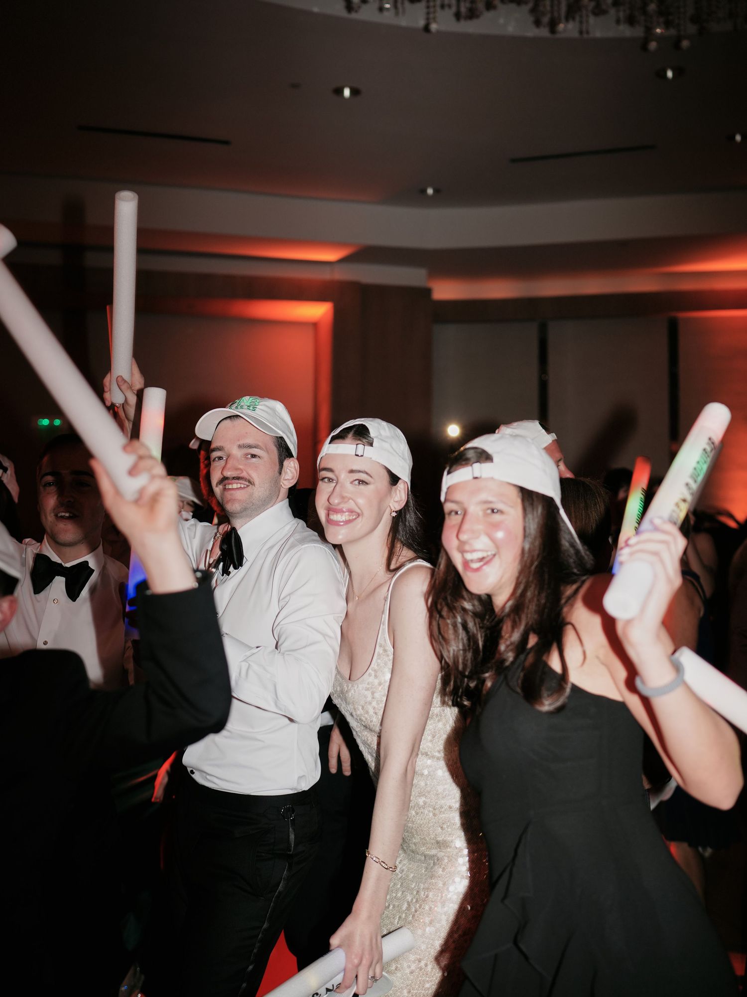 Party guests dance and celebrate with glowing LED foam sticks in a reception hall with red ambient lighting.
