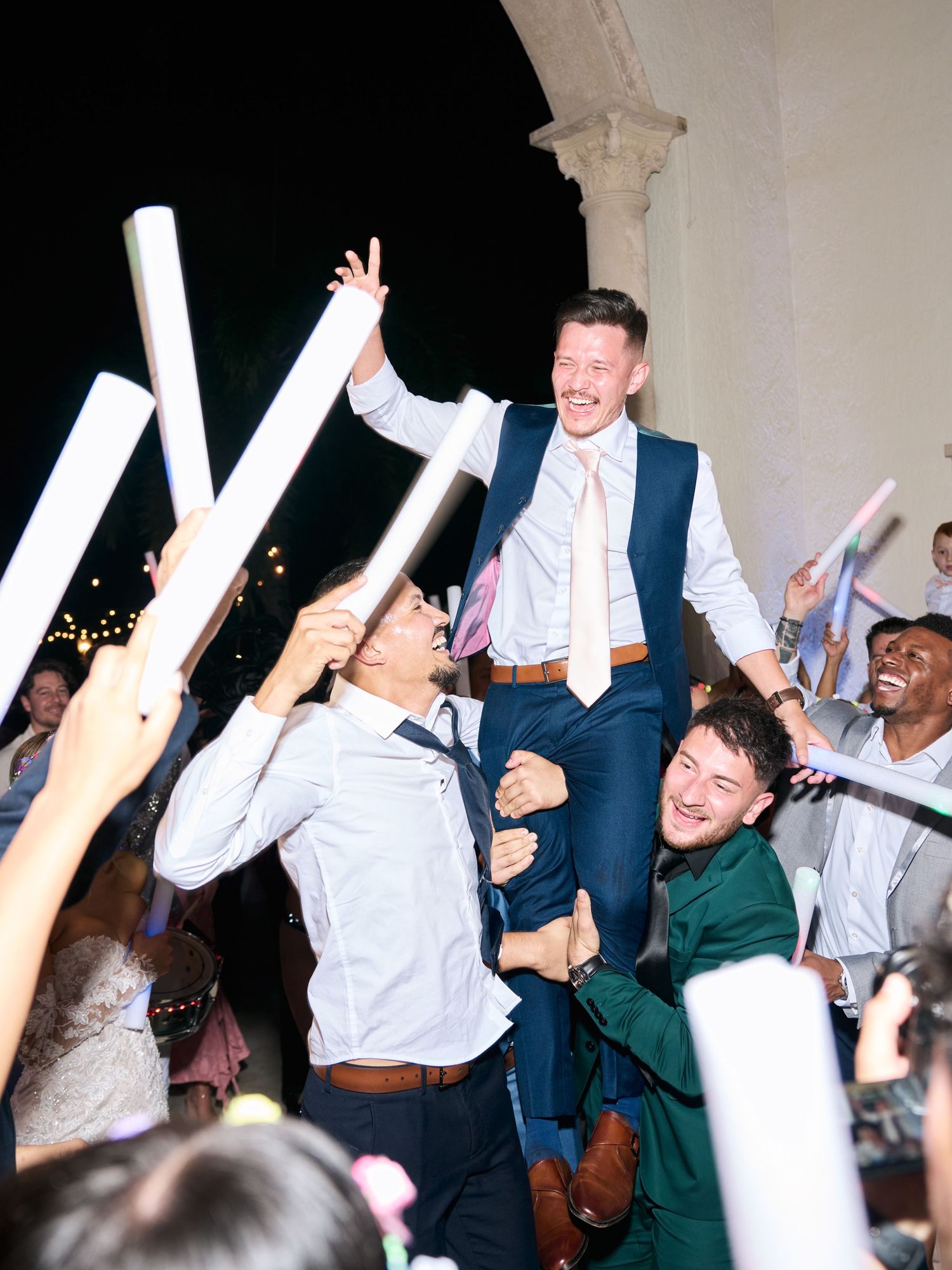 Groom crowd surfing during lively wedding reception celebration with guests holding light wands.