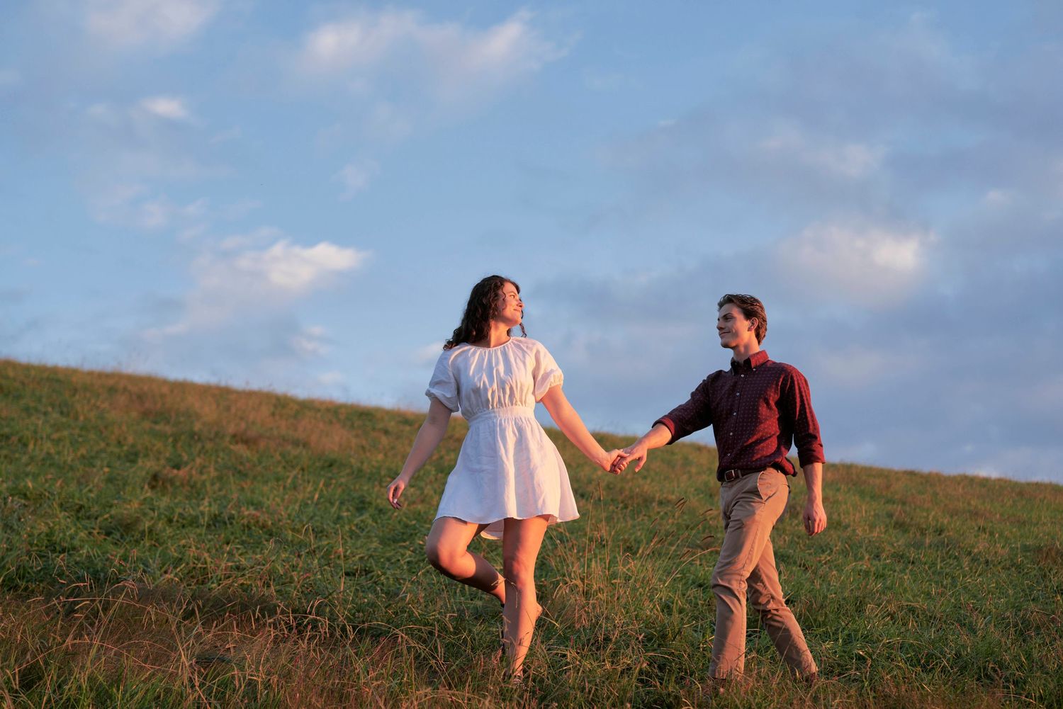 Two people hold hands while running together on a grassy hill at sunset.