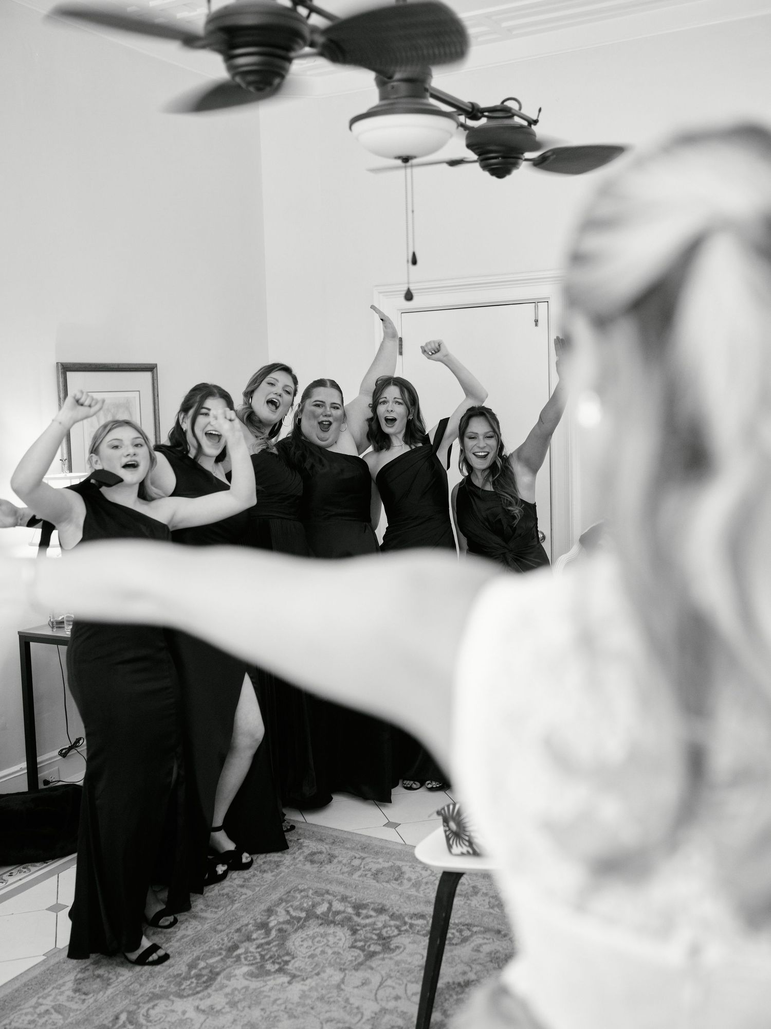 Black and white photo of a bride revealing her wedding dress to excited bridesmaids in black gowns cheering with joy.