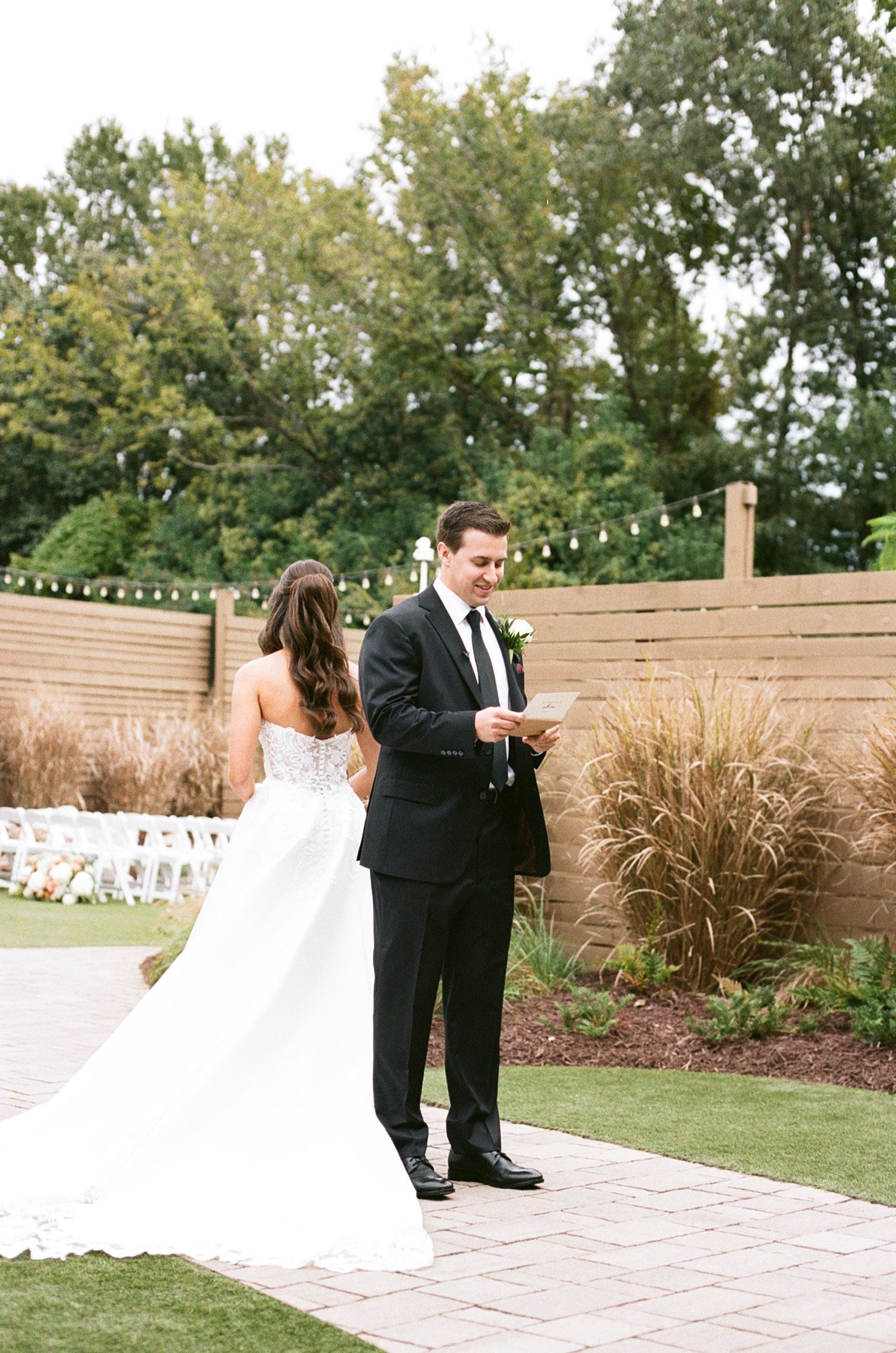 Groom reads vows to bride in strapless white gown during outdoor wedding ceremony with string lights and lush greenery.