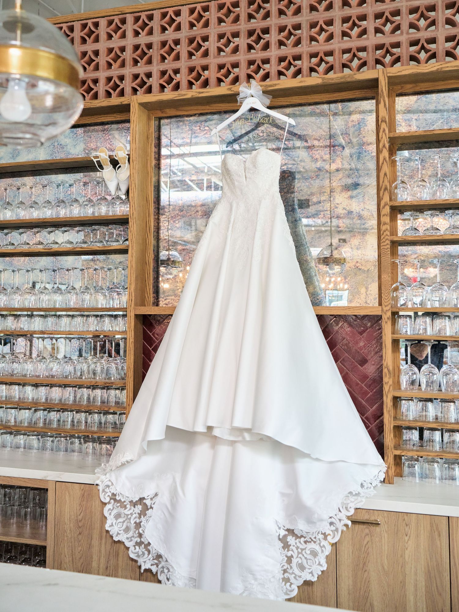 White strapless wedding gown with lace trim hem hanging in a wine bar with glass-lined wooden shelves.